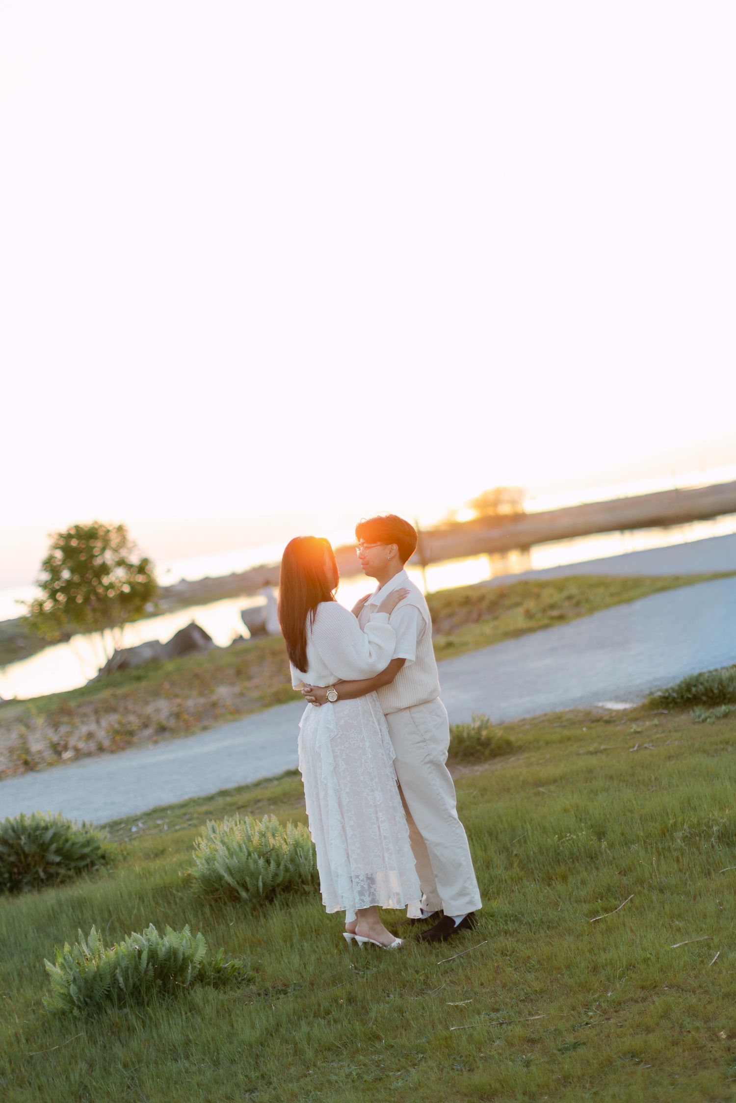 Couple in white and cream outfits embrace on a grassy lawn by the water as the sun sets behind them.