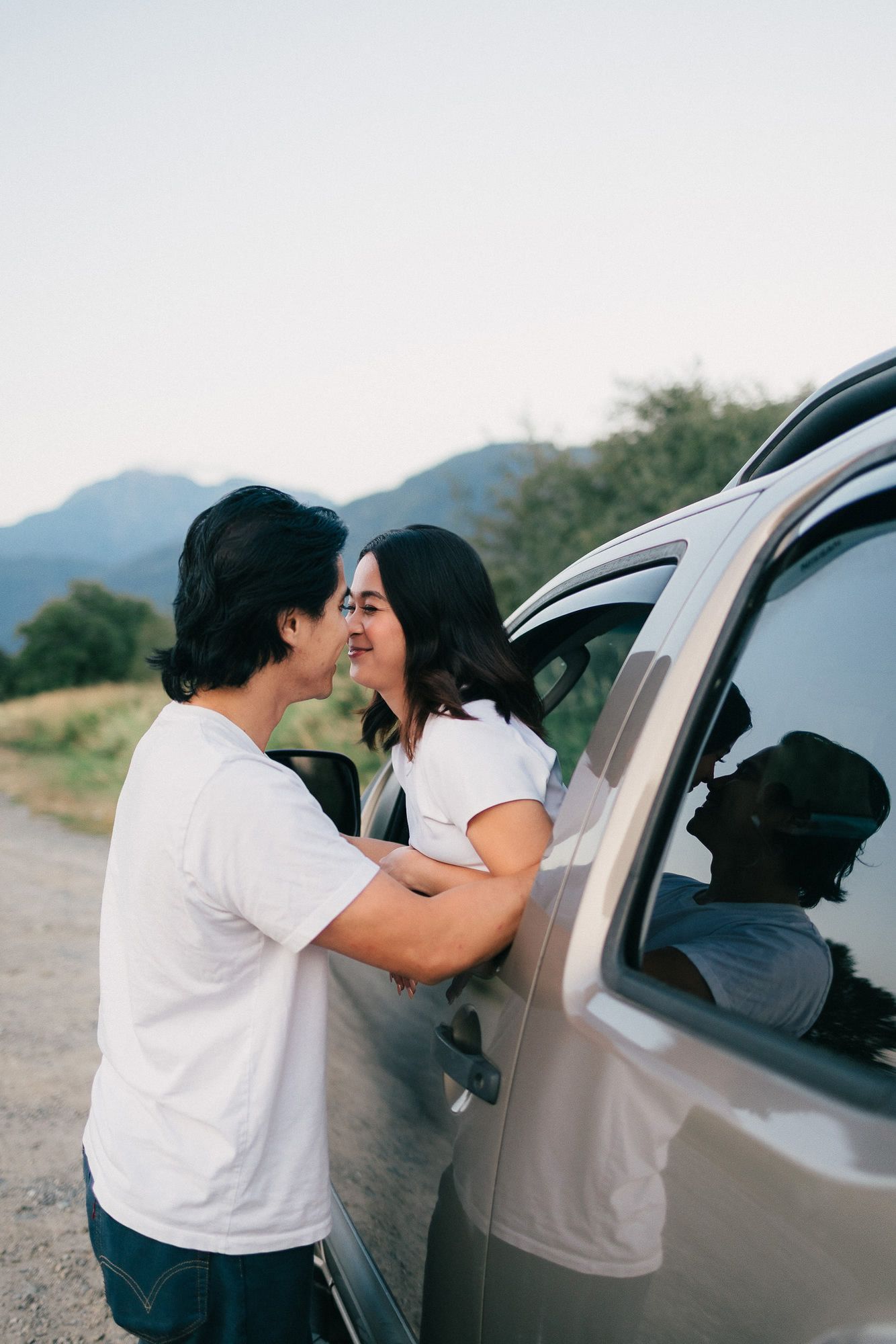 Couple in white t-shirts share a tender moment as a woman leans out of a car window to kiss a man, with mountains behind them.