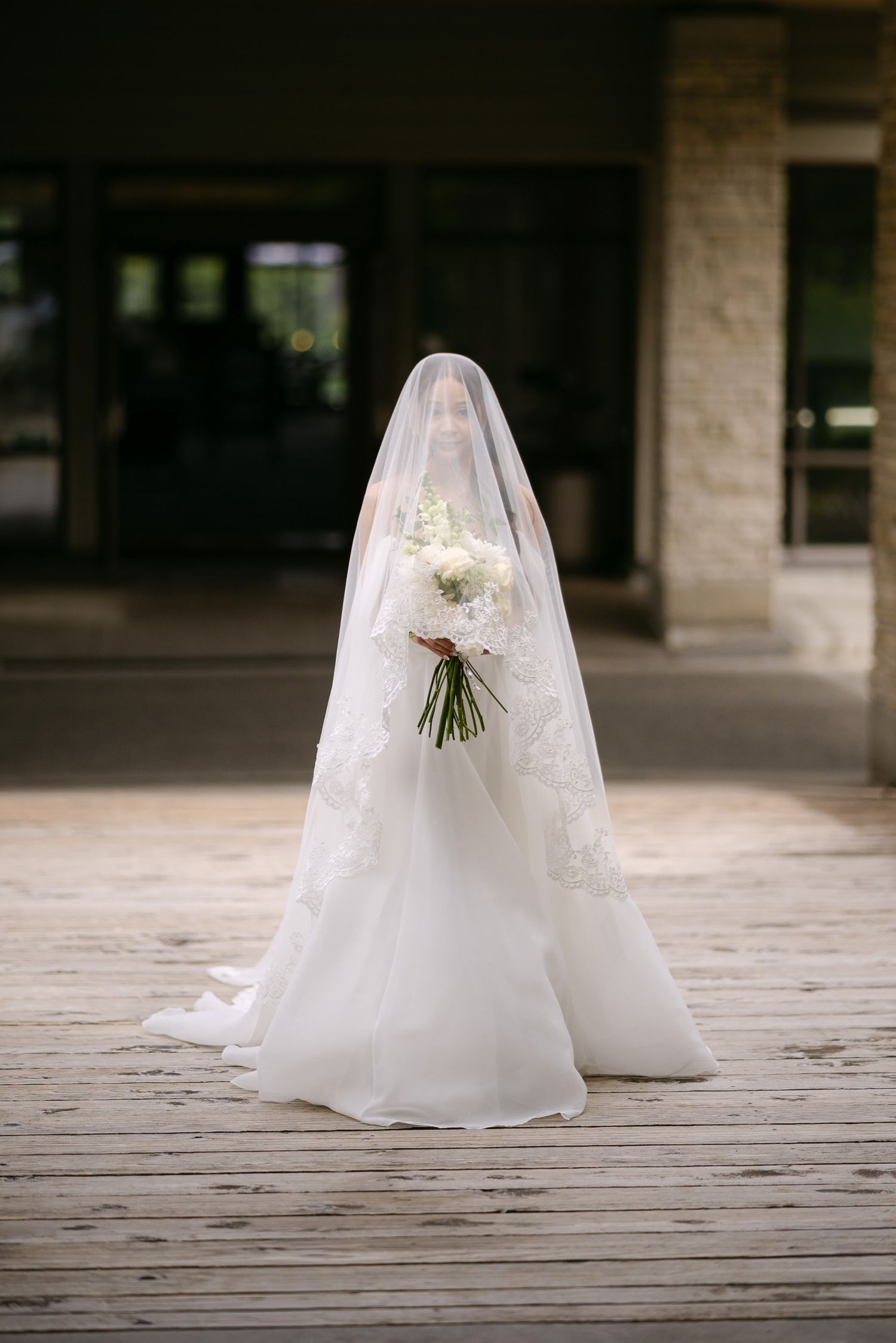 Bride in a flowing white wedding gown and lace veil holding a white floral bouquet on a wooden deck.