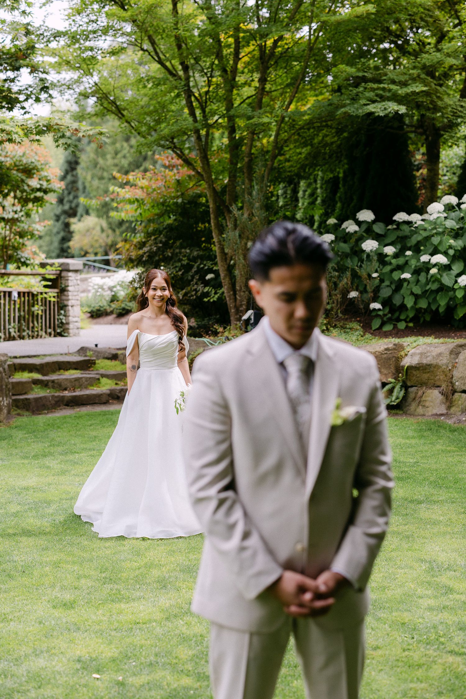 Bride in white gown approaches groom in tan suit during outdoor first look in lush garden setting.