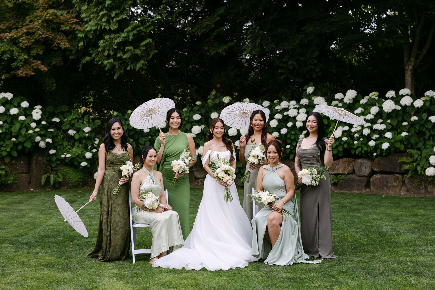 Bride in white gown poses with six bridesmaids in green and gray dresses holding parasols and bouquets outdoors.