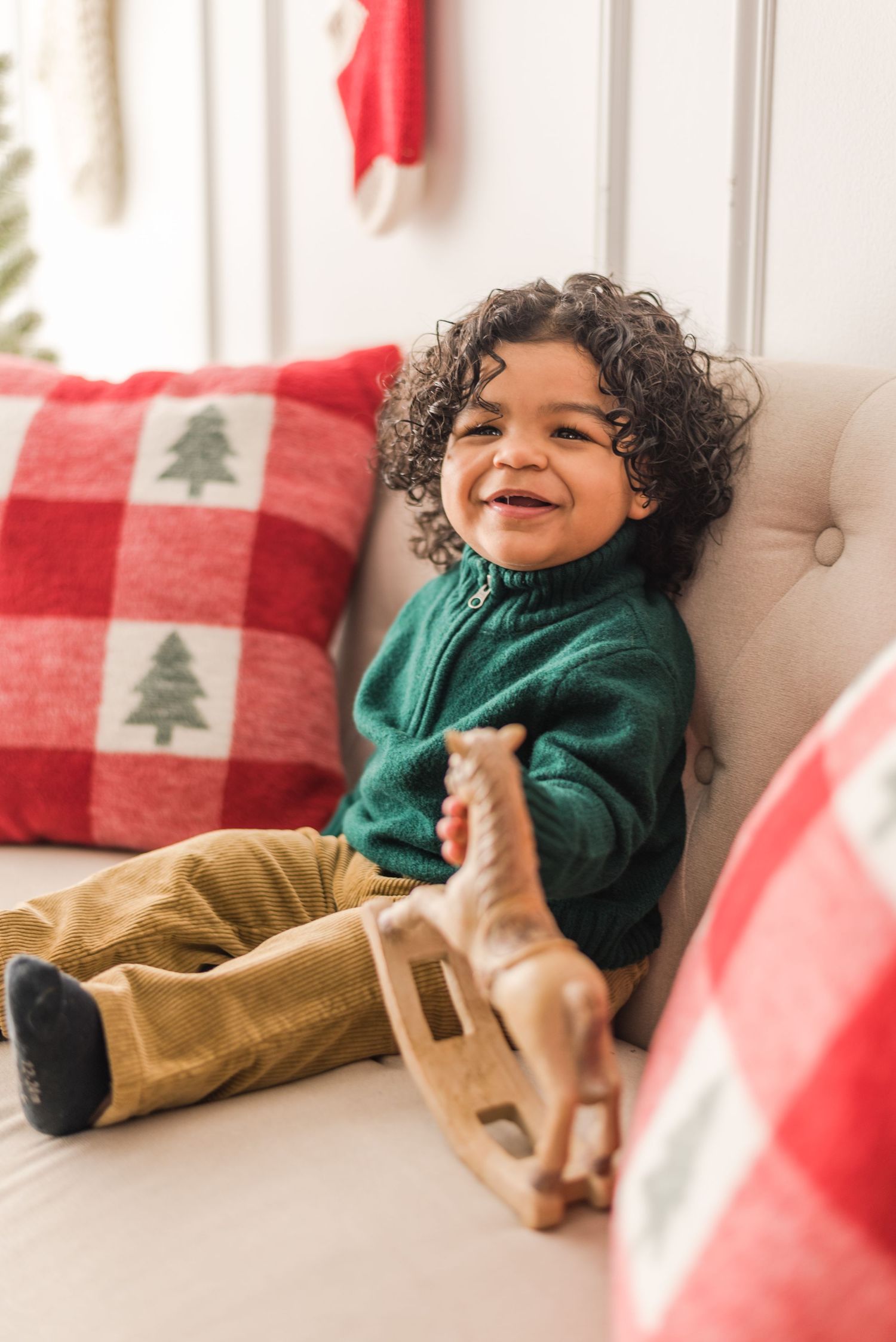 Toddler in green sweater and brown pants sits on white chair with festive red plaid Christmas pillows and wooden toy.
