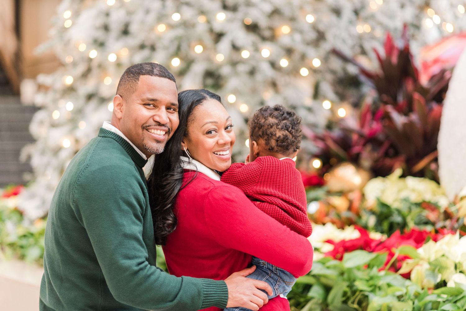 Happy family wearing green and red sweaters poses together in front of holiday decor at a garden center.