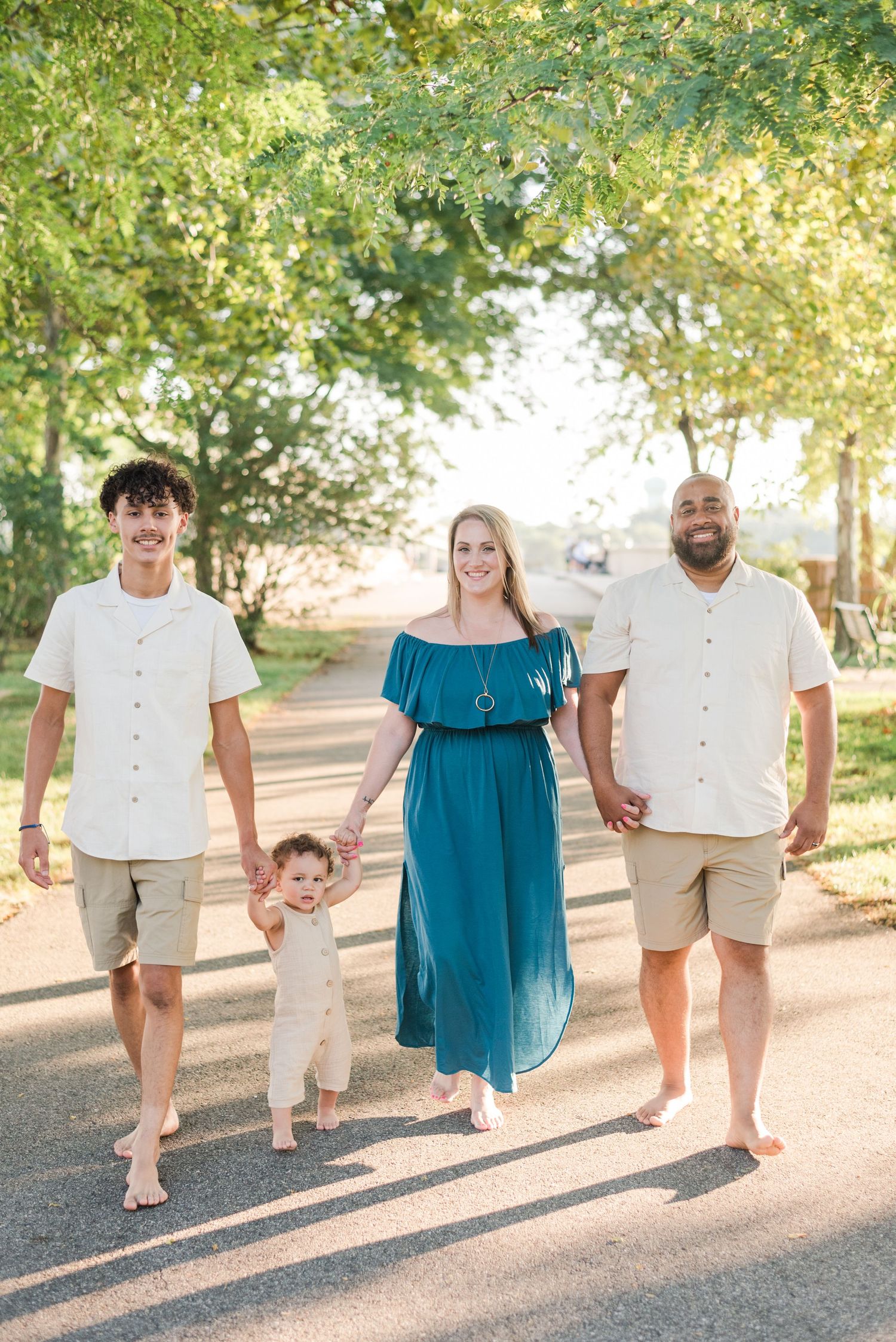 A group walks together on a sunlit path under trees, casting long shadows while wearing casual summer attire.
