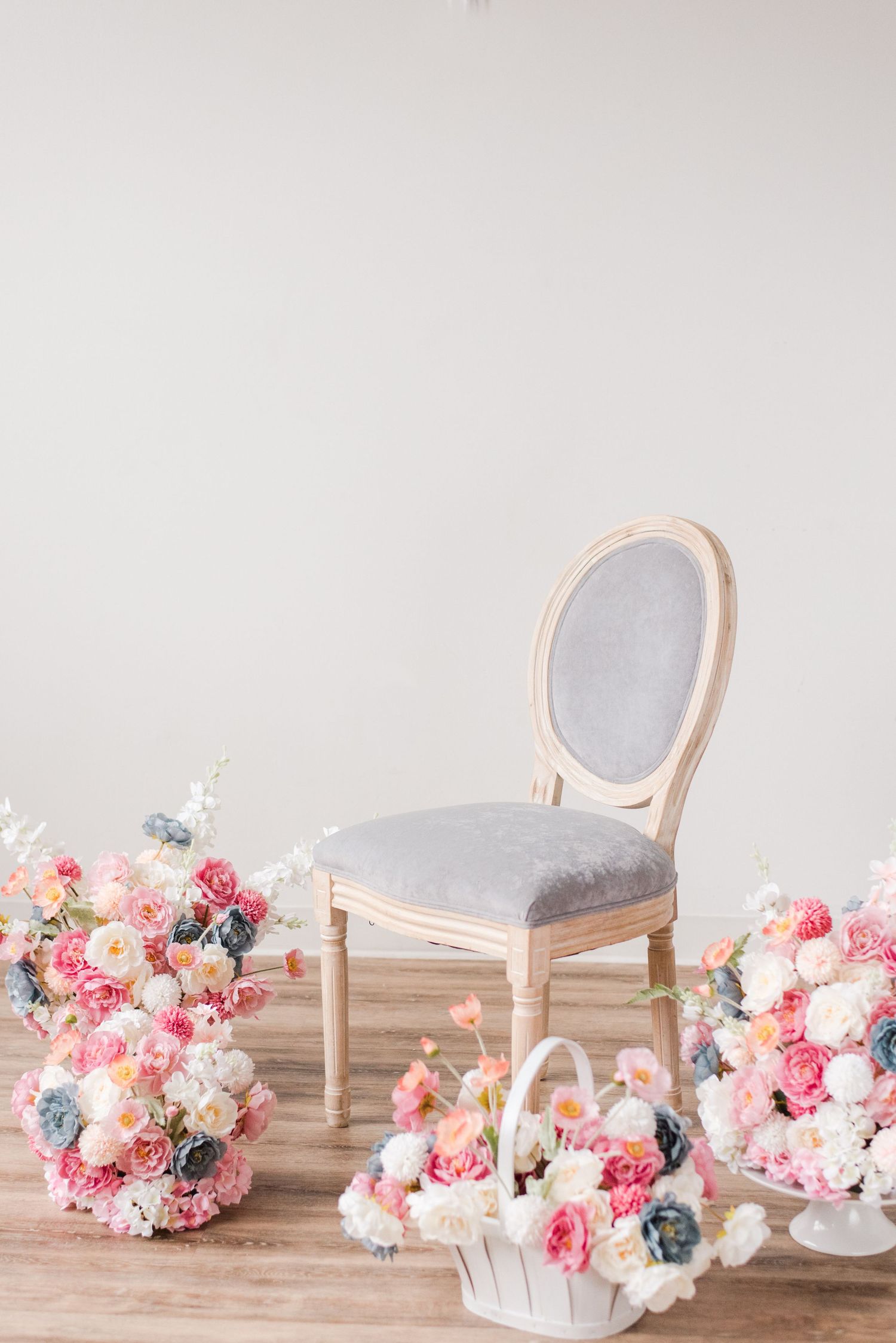 Elegant French provincial chair with grey velvet upholstery surrounded by pink and white floral arrangements on wooden floor.