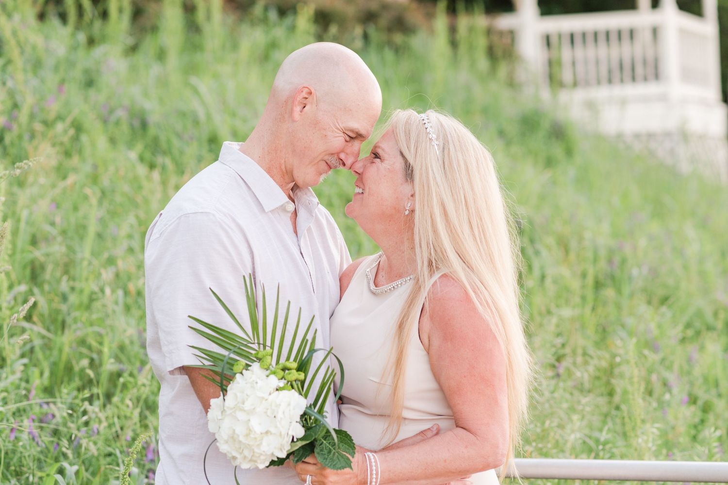A romantic couple shares an intimate moment in a grassy field while holding white flowers.