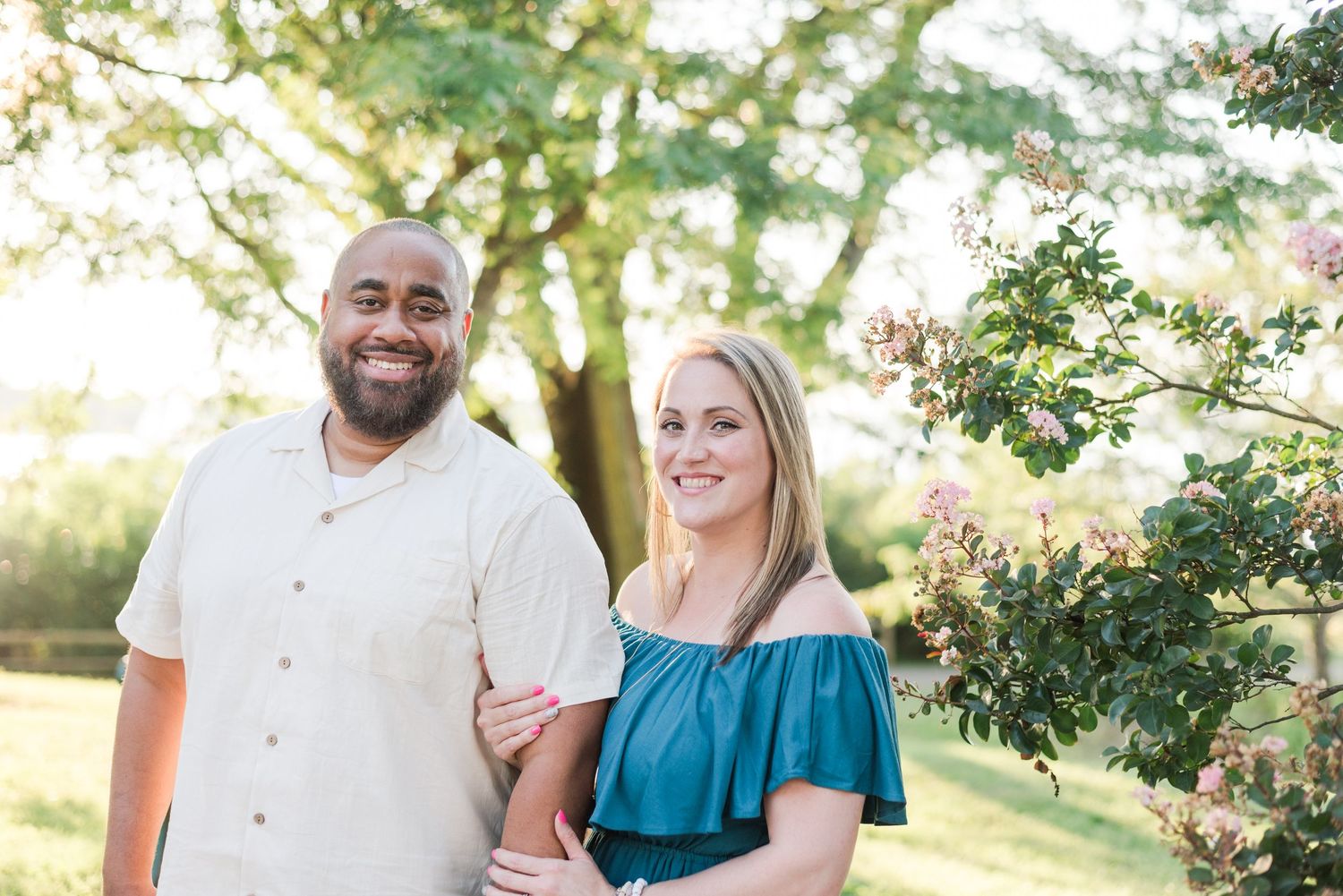 A couple poses outdoors in casual attire with sunlight filtering through trees during a summer photo session.