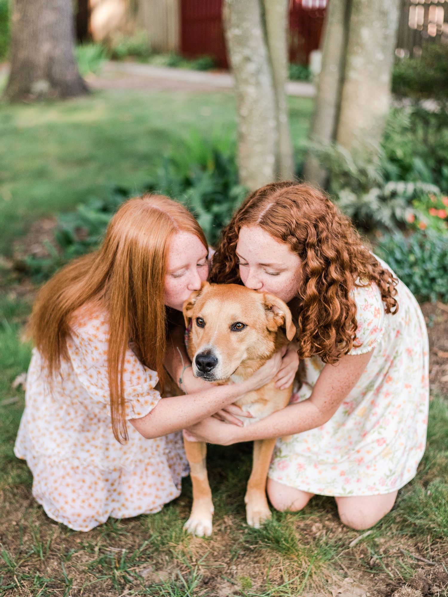 Two people in floral dresses share an affectionate moment with their golden mixed breed dog in a garden setting.