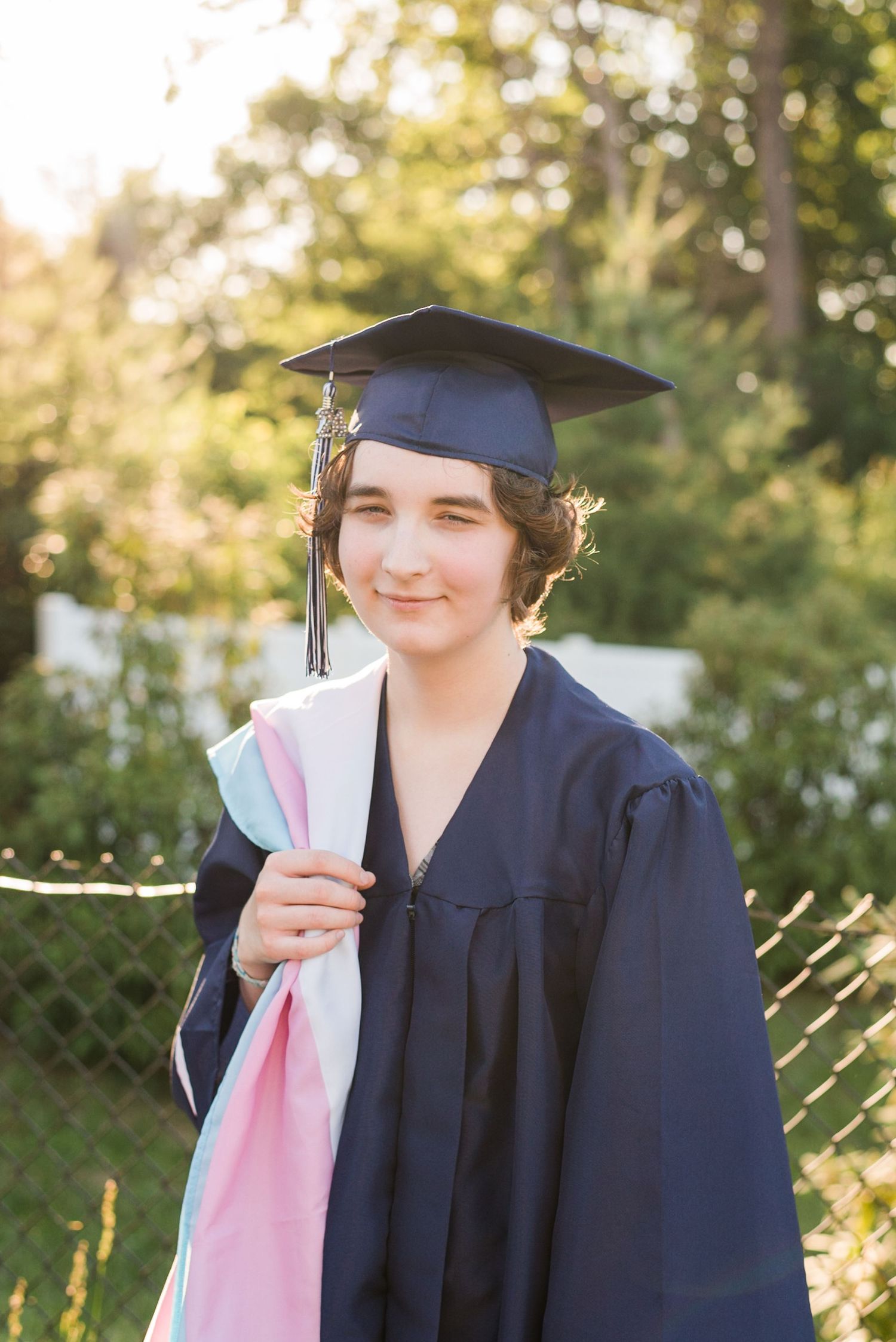 Graduate in navy cap and gown holding diploma while smiling in sunny outdoor setting during commencement ceremony.