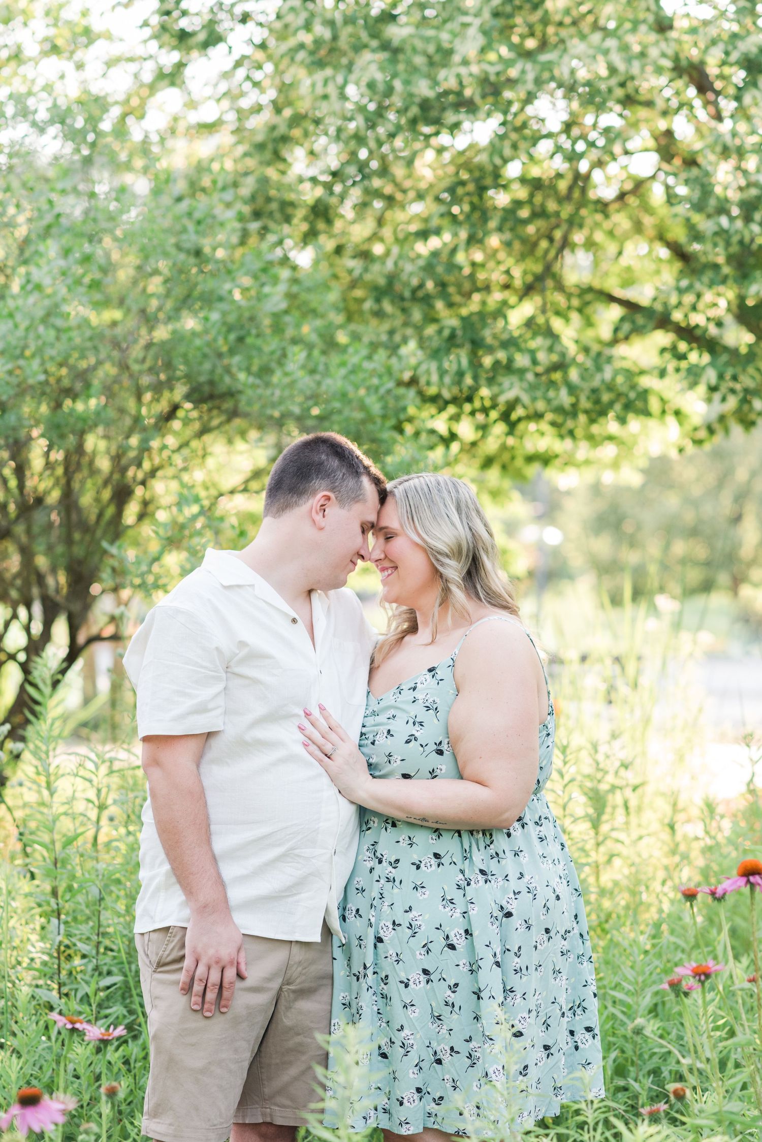Couple shares intimate moments in a sunny meadow with wildflowers and trees during golden hour.