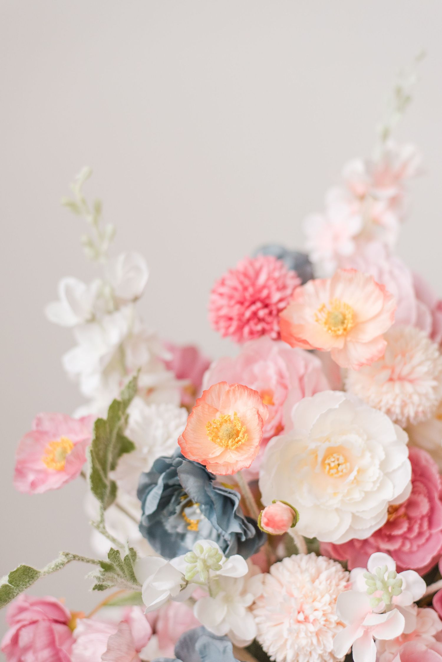 Soft romantic floral arrangement with pink carnations, white roses, and dusty blue flowers against a light gray background.