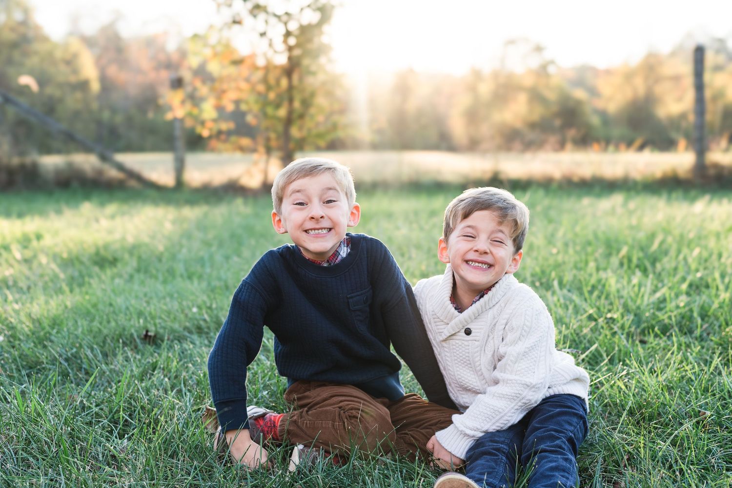 Two people laugh together while sitting in a green grassy field during golden hour sunlight.