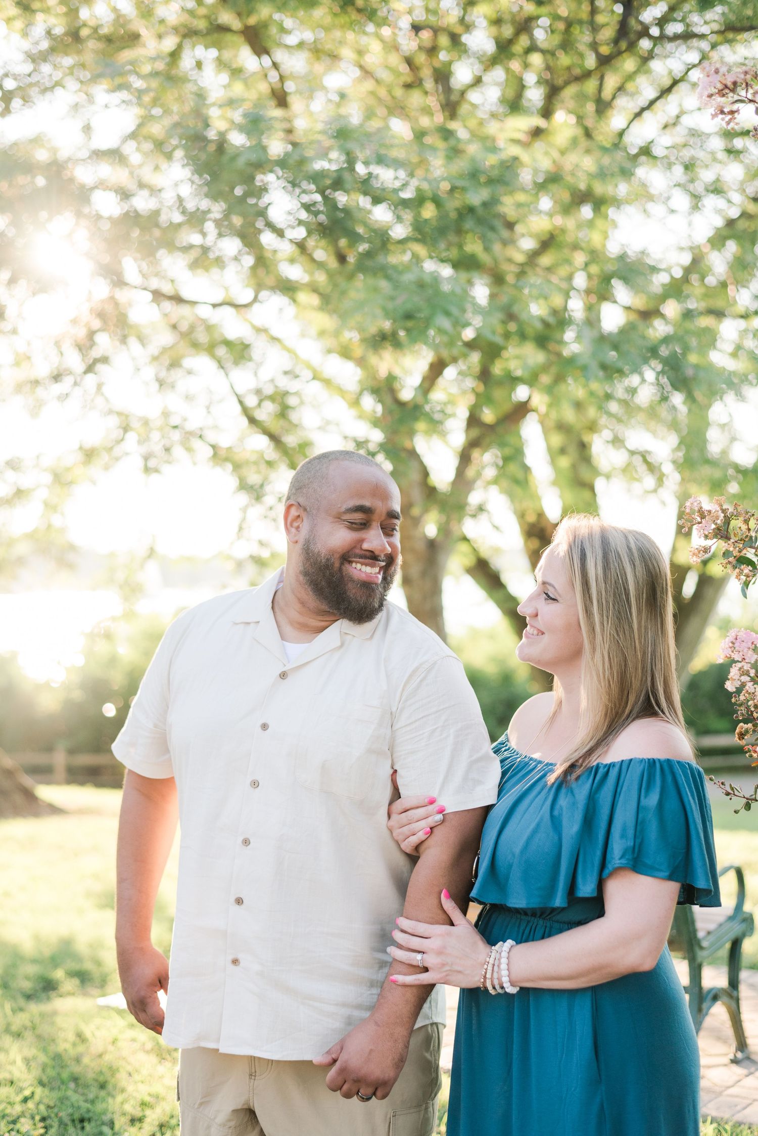 A couple shares a sweet moment outdoors with golden sunlight streaming through green trees on a summer evening.