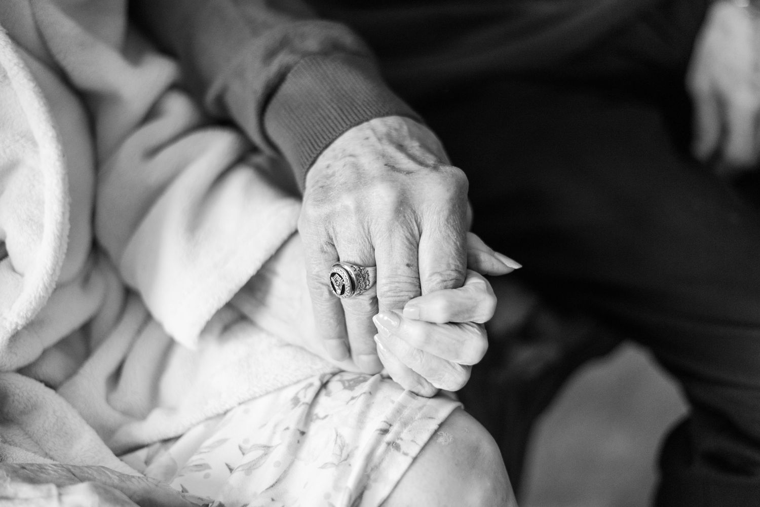 Close up black and white photo of an elderly person's hands with a ring resting on their lap.