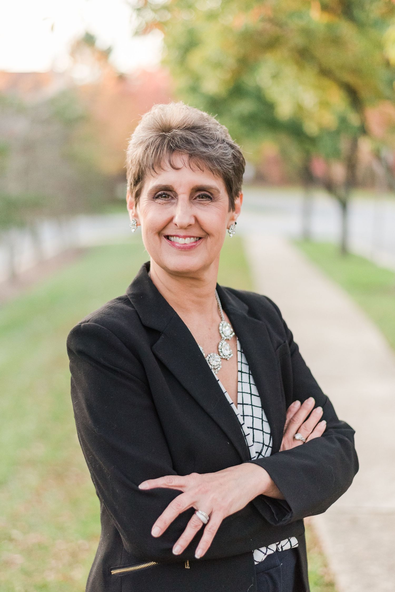 Professional headshot of someone in a black blazer and patterned blouse outdoors on a tree-lined path.