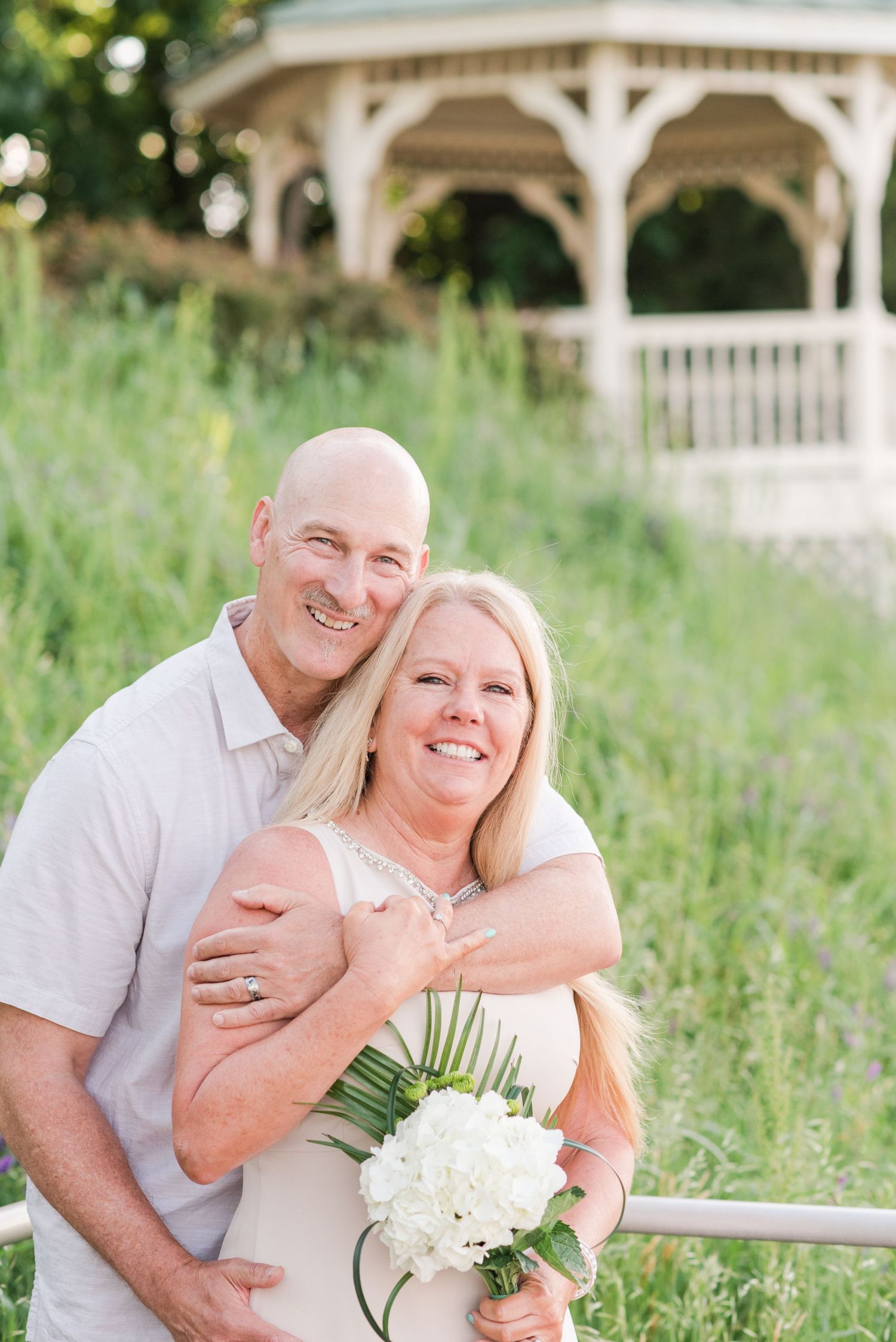 Happy couple embrace outdoors in front of white gazebo with bride holding white bouquet in summer garden setting.