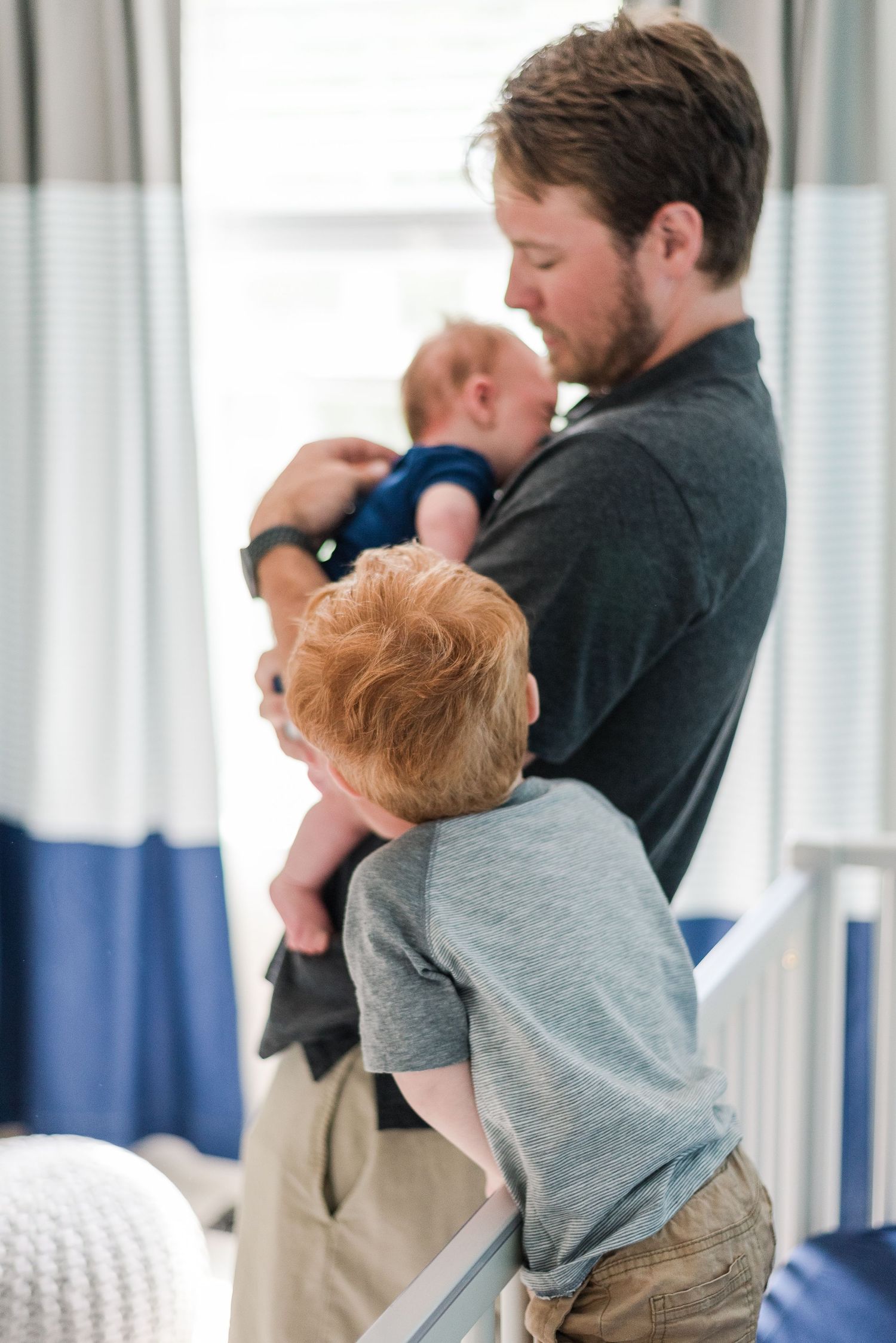 A father holding an infant while another child in a gray t-shirt hugs his leg, standing in front of sheer curtains.