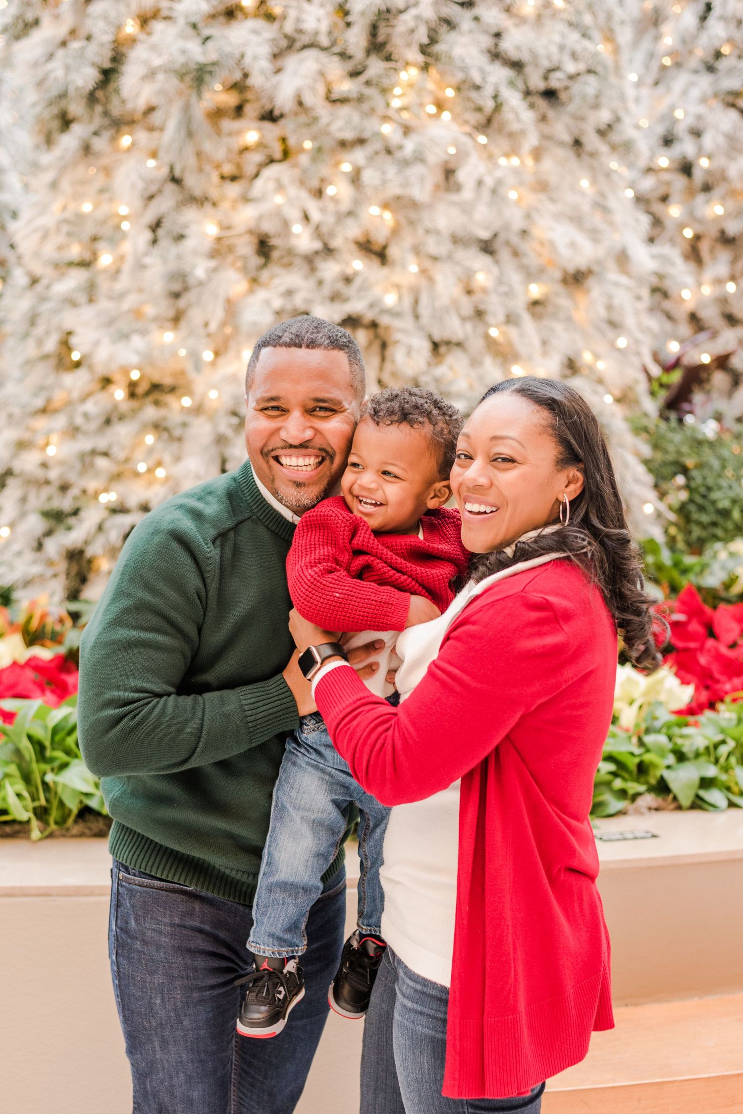 A family poses together outdoors during winter, wearing festive red and green sweaters with poinsettias in the background.