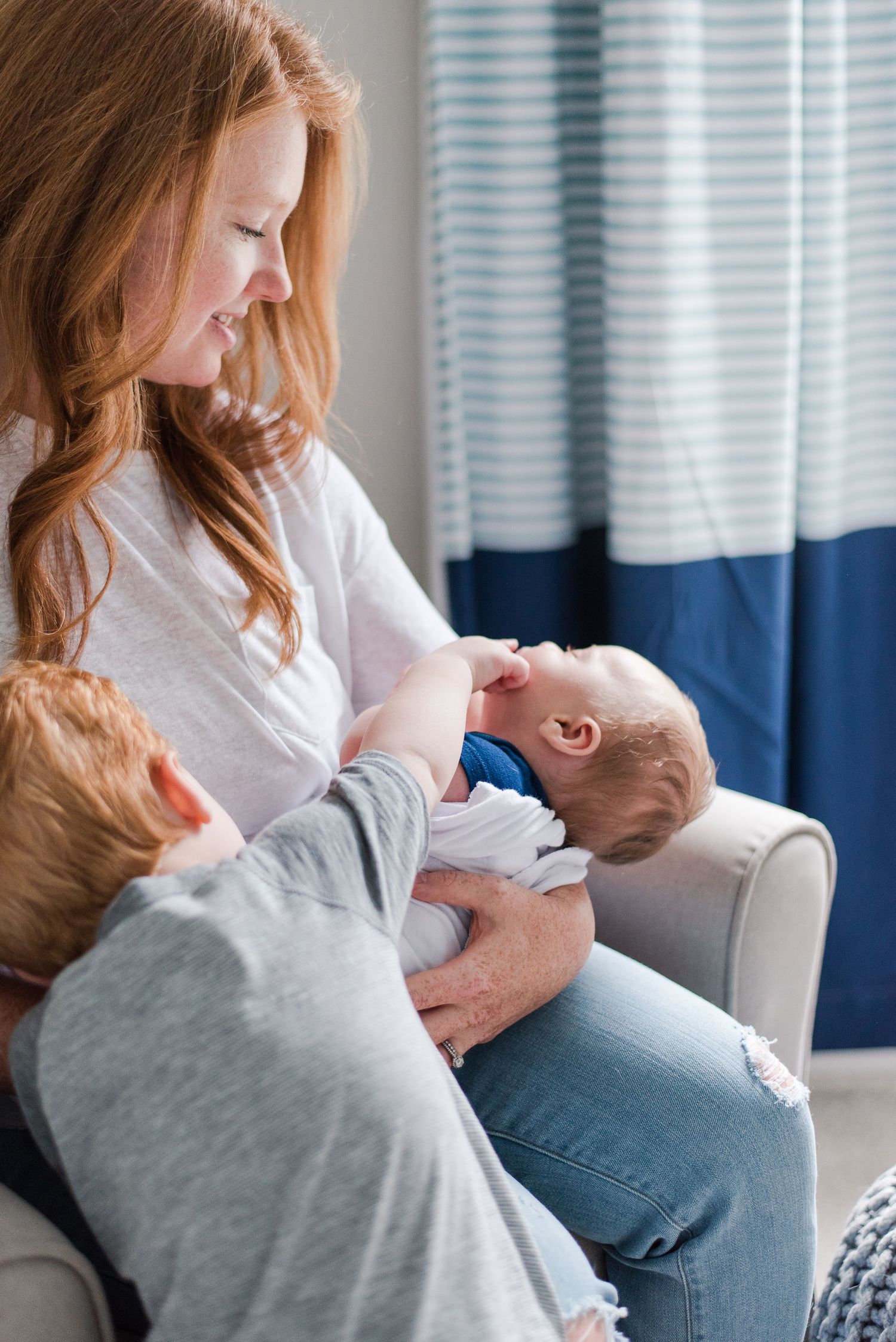 Parent sits in armchair holding and feeding an infant while wearing casual clothing next to blue and white curtains.