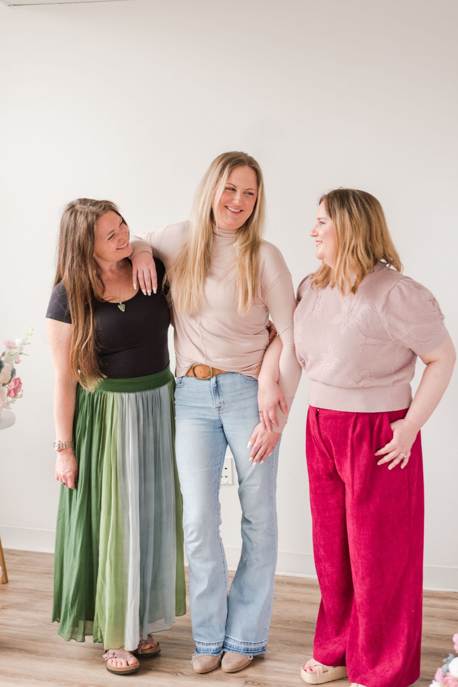 Three friends laughing together in a bright studio wearing contrasting outfits of green, denim and pink.