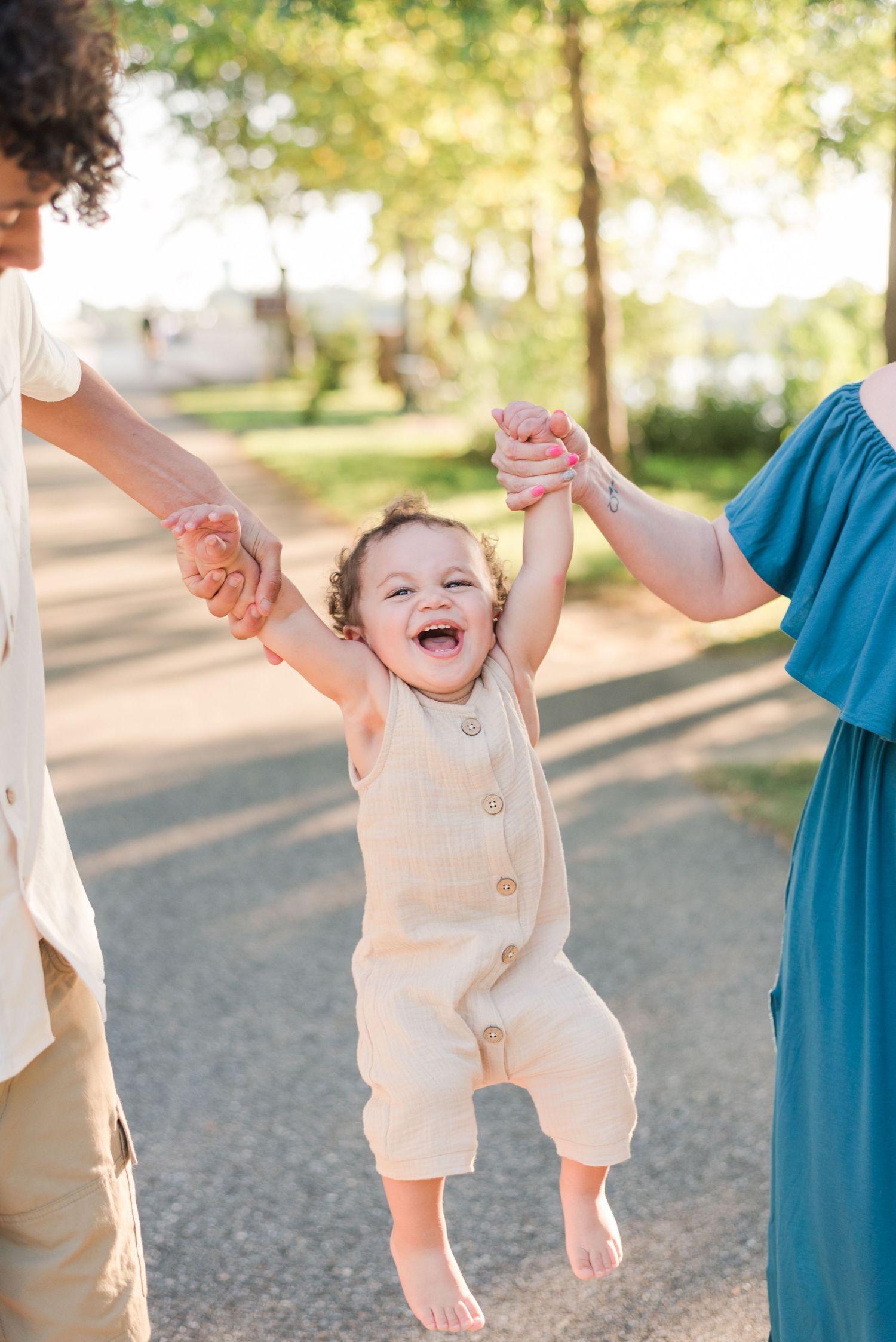 Laughing baby in cream-colored romper being lifted by arms in a sunny park setting during golden hour.