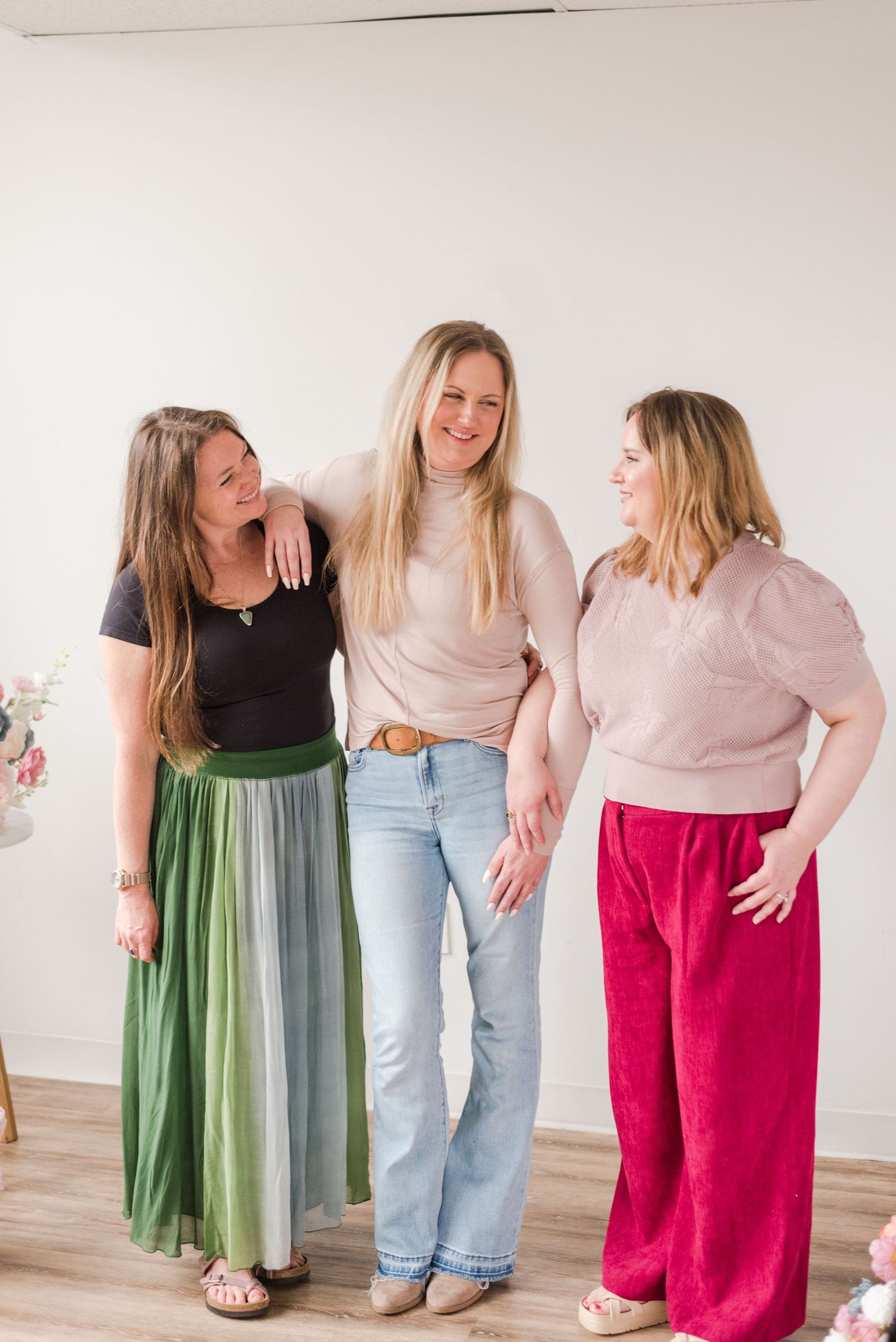 Three friends sharing a candid moment together, wearing casual outfits including a green skirt, jeans, and red pants.