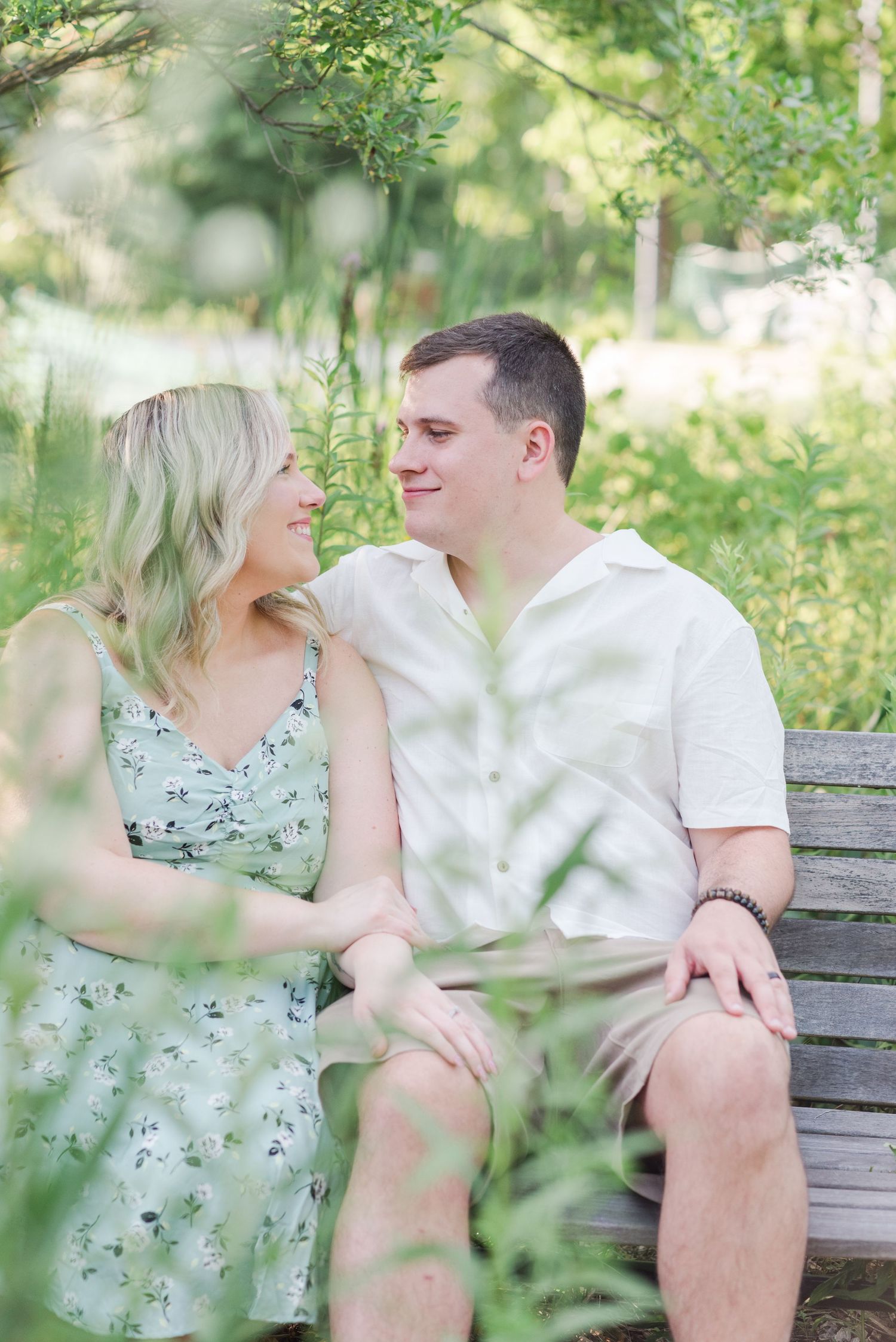 A couple shares a romantic moment while sitting on a wooden bench surrounded by lush green foliage in a garden setting.