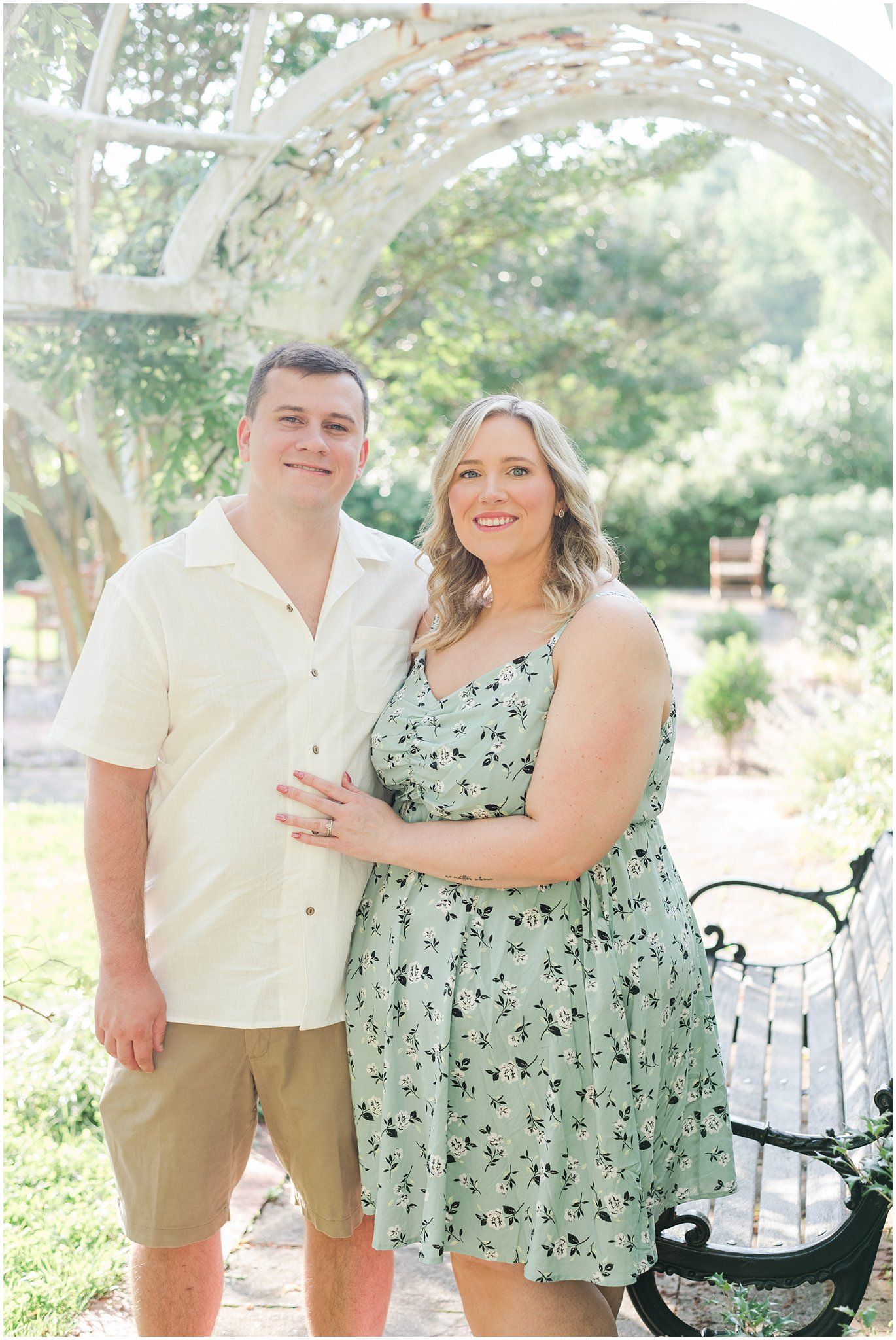 A loving couple in matching light colored outfits poses together on a garden path during a sunny summer photoshoot.