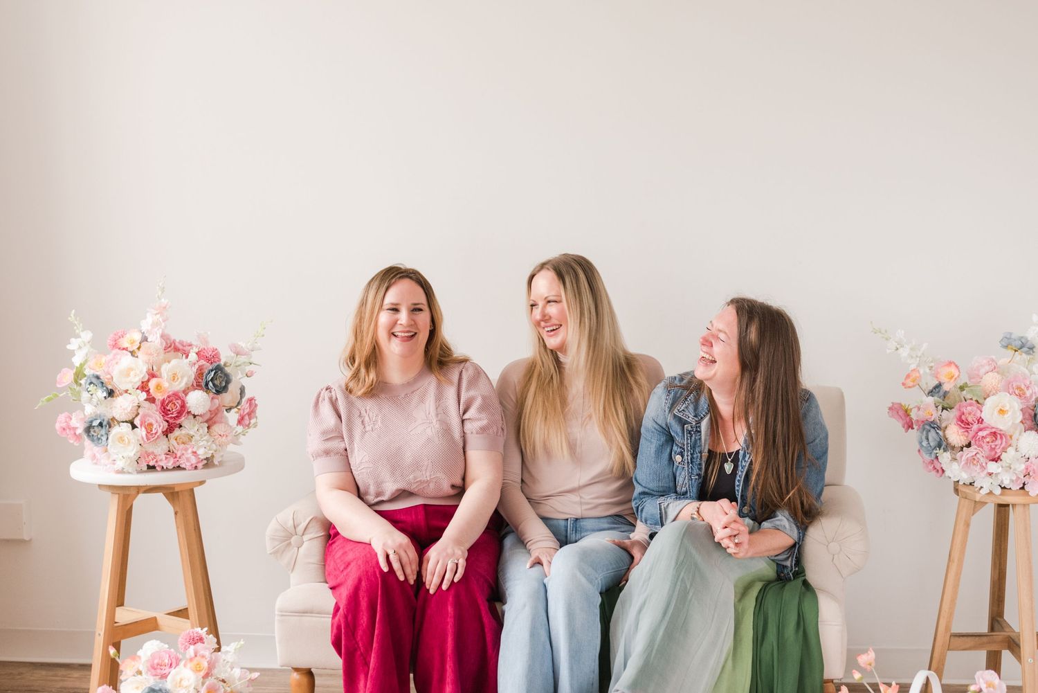 Three friends sit casually on a white bench between decorative floral arrangements in a bright studio setting.