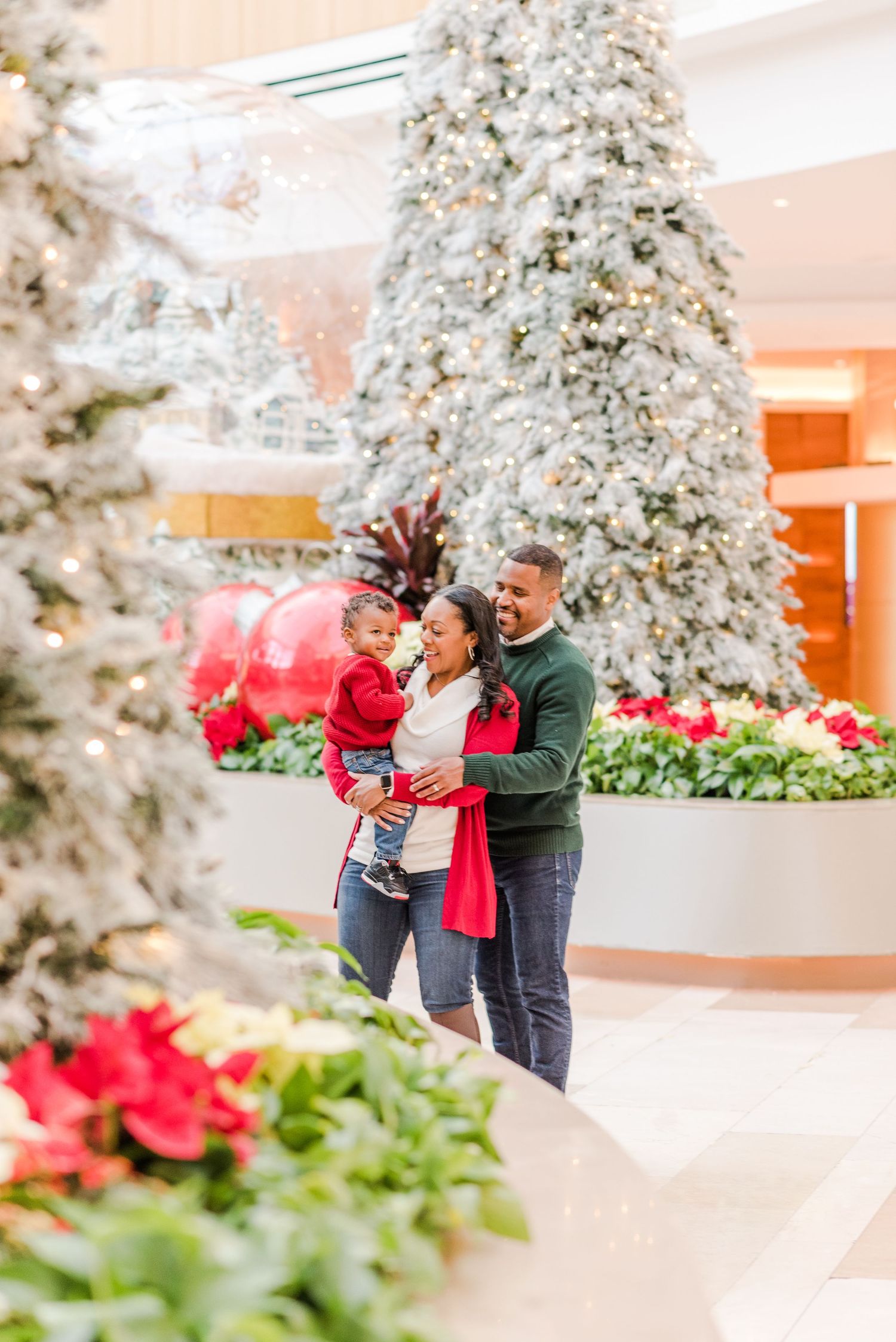 A family admires towering flocked Christmas trees and red poinsettias in a brightly lit indoor holiday display.