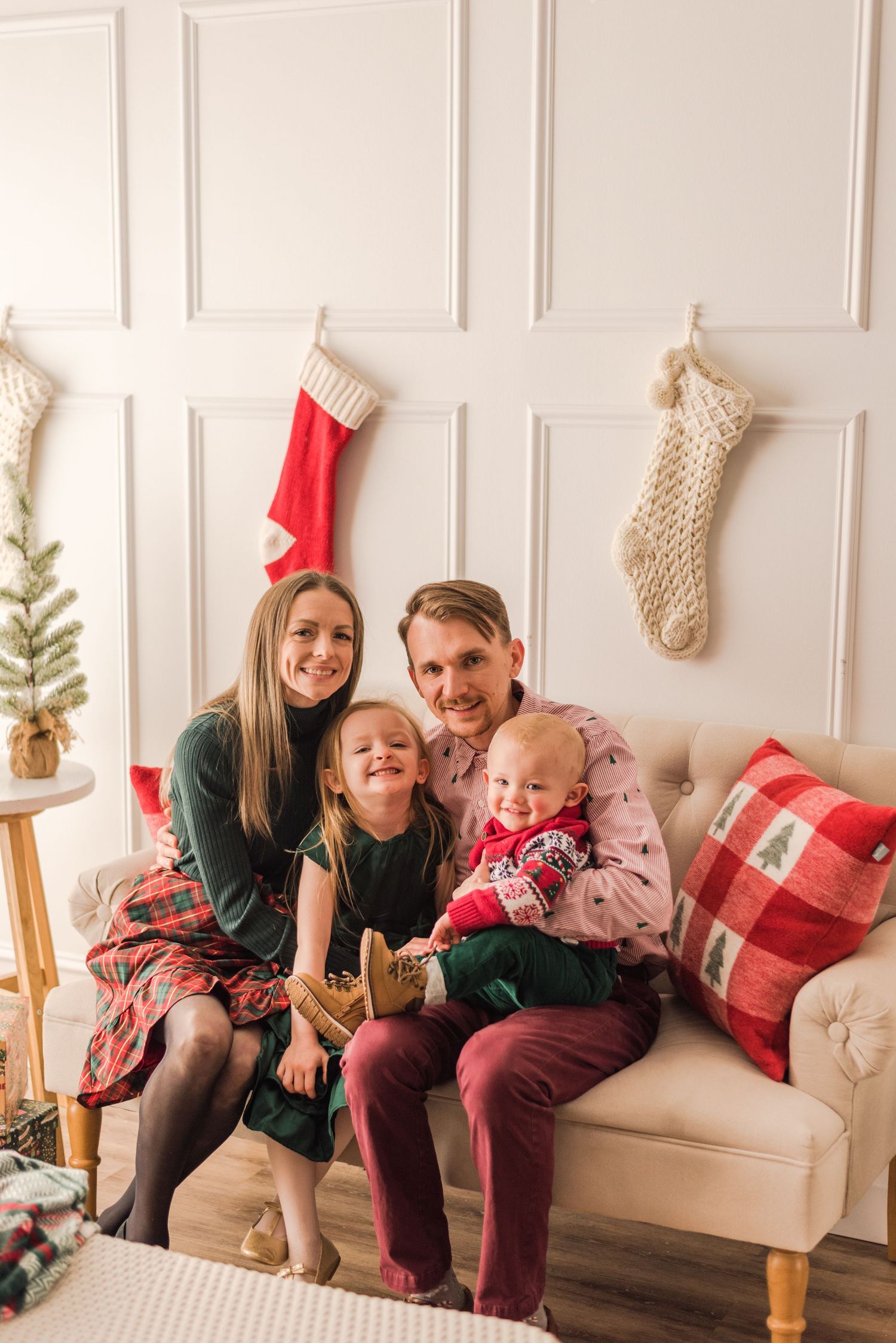 Family sits together on white bench displaying Christmas stockings and decorations on wall behind them.