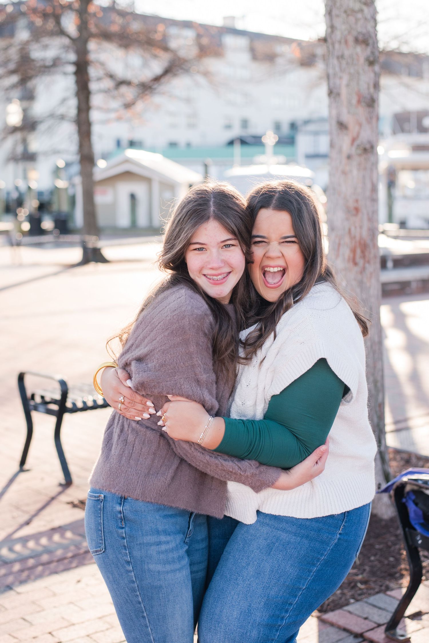 Two friends share a joyful embrace and laugh together on a sunny day in an outdoor plaza setting.