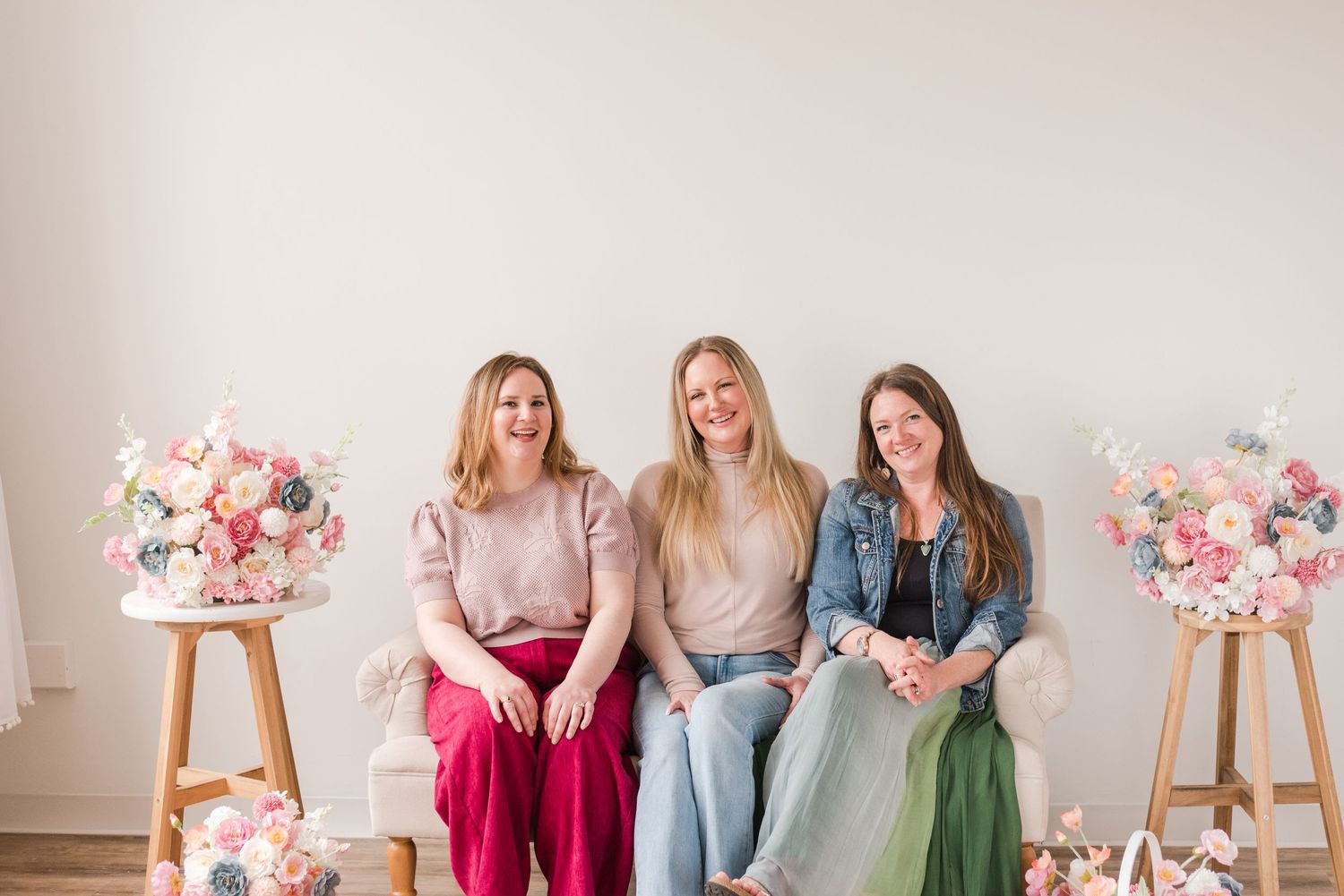 Three women sitting together on a bench with floral arrangements on wooden easels beside them against a neutral wall.