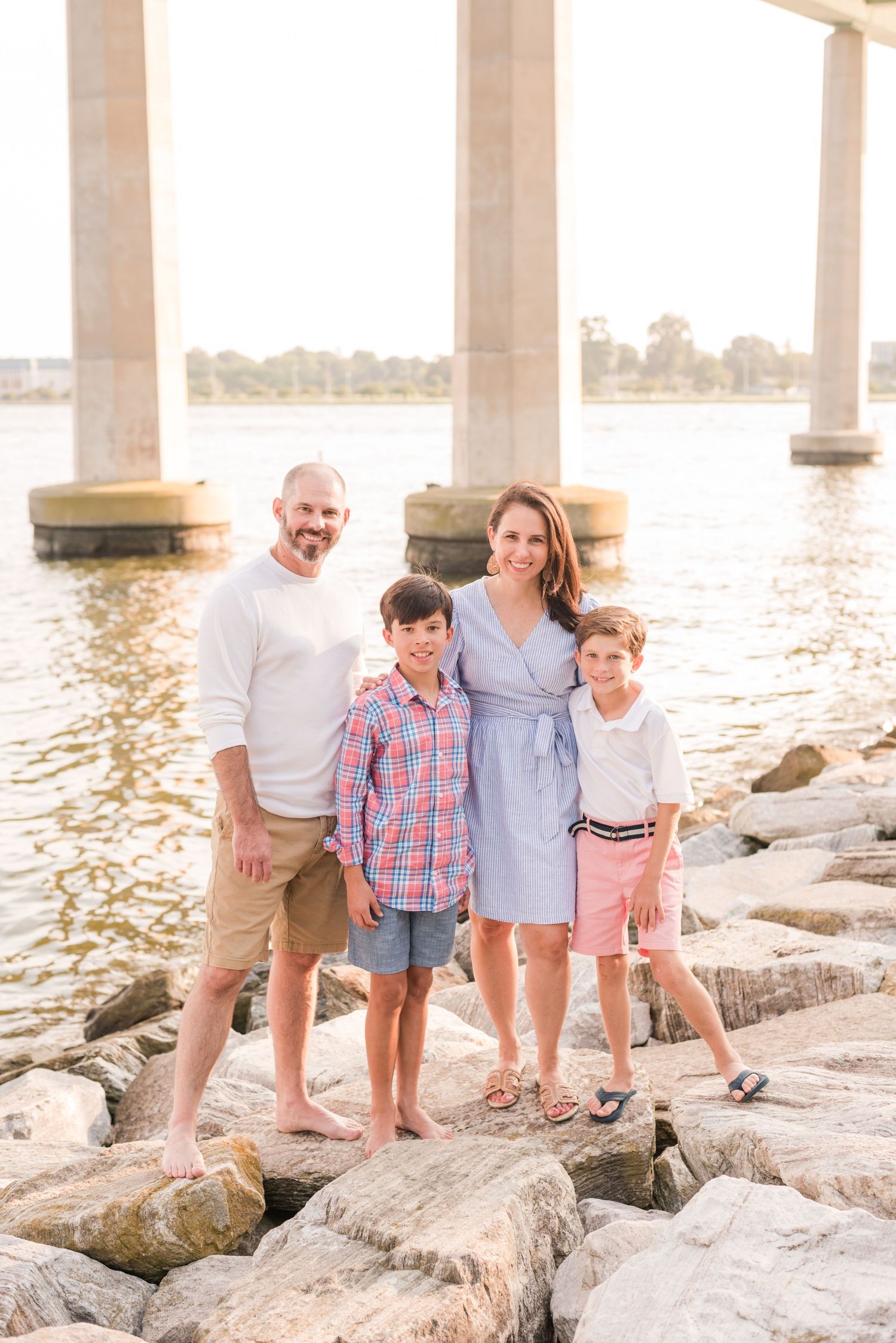 Family standing together on rocky shoreline under bridge at sunset with water in background.