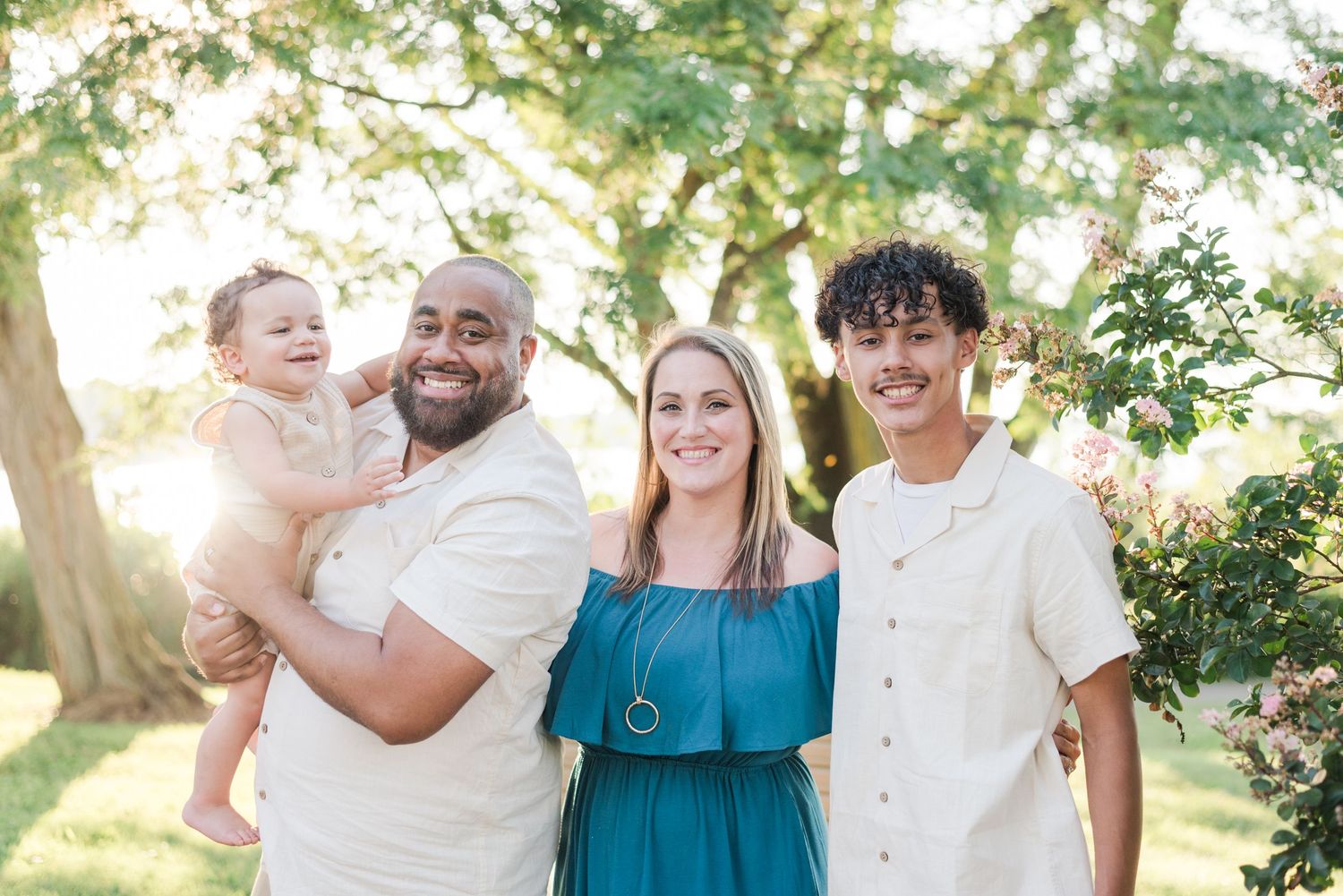 A loving family stands together outdoors in warm sunlight with trees providing a natural backdrop.