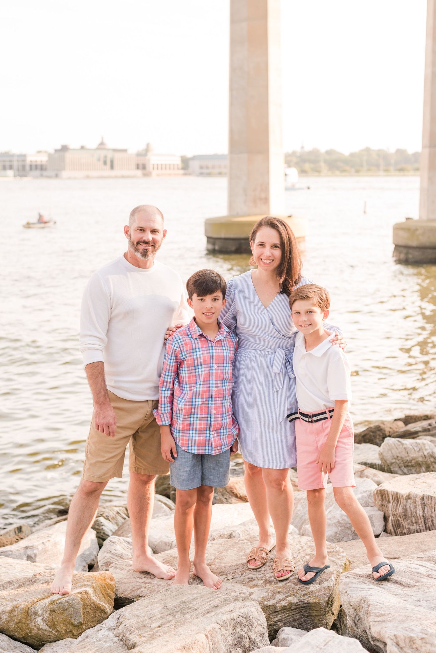 Happy family poses together on coastal rocks at sunrise with water and buildings in background.