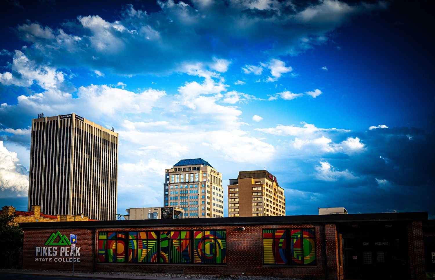 Pikes Peak State College building with colorful graffiti mural beneath a dramatic blue sky and downtown Colorado Springs skyline.