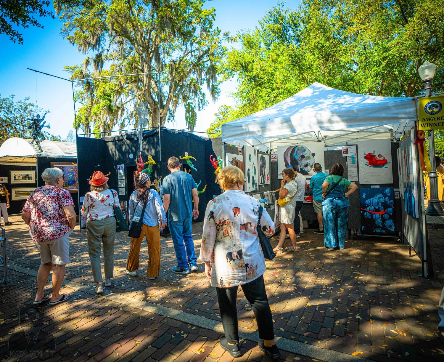 Outdoor art festival with white tents and visitors browsing artwork displays on a sunny day.