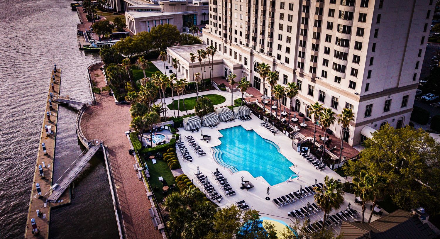 Aerial view of a luxurious hotel swimming pool with lounge chairs surrounded by high-rise buildings along a waterfront.