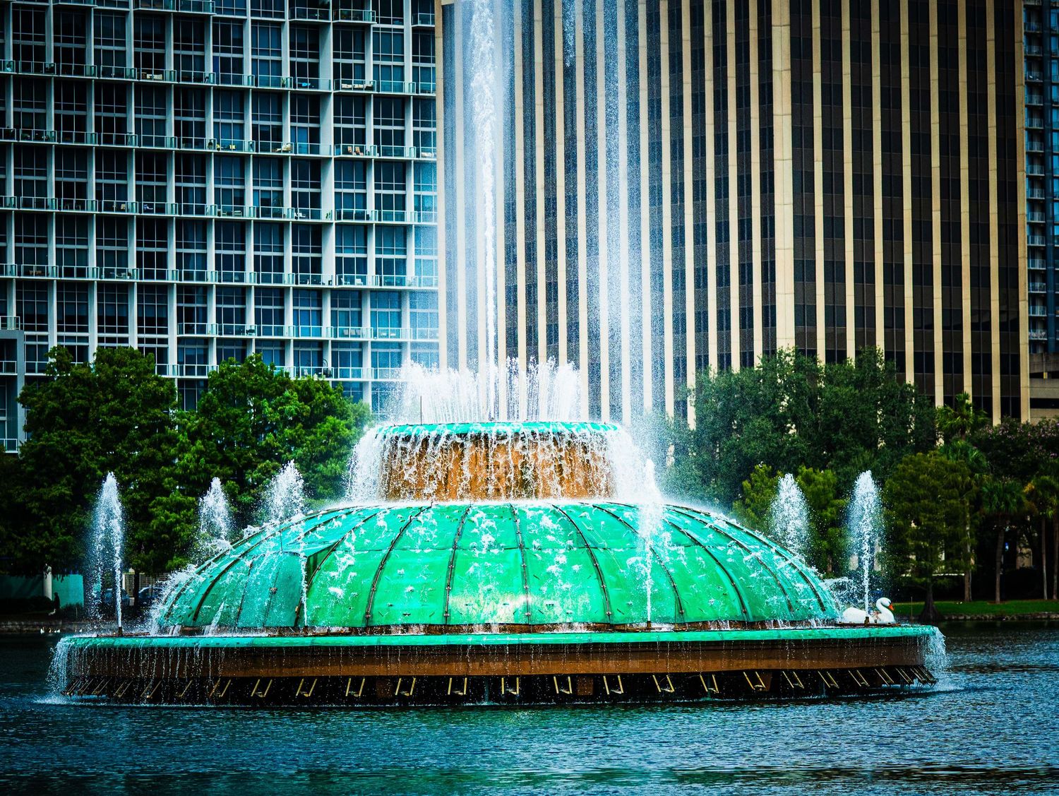 Close-up of Orlando's iconic green Lake Eola fountain with multiple water jets and city buildings in background.