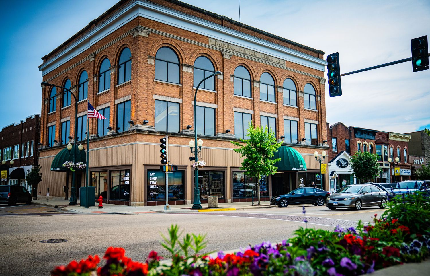 Historic brick buildings and colorful flowers line a small town main street under bright blue skies.