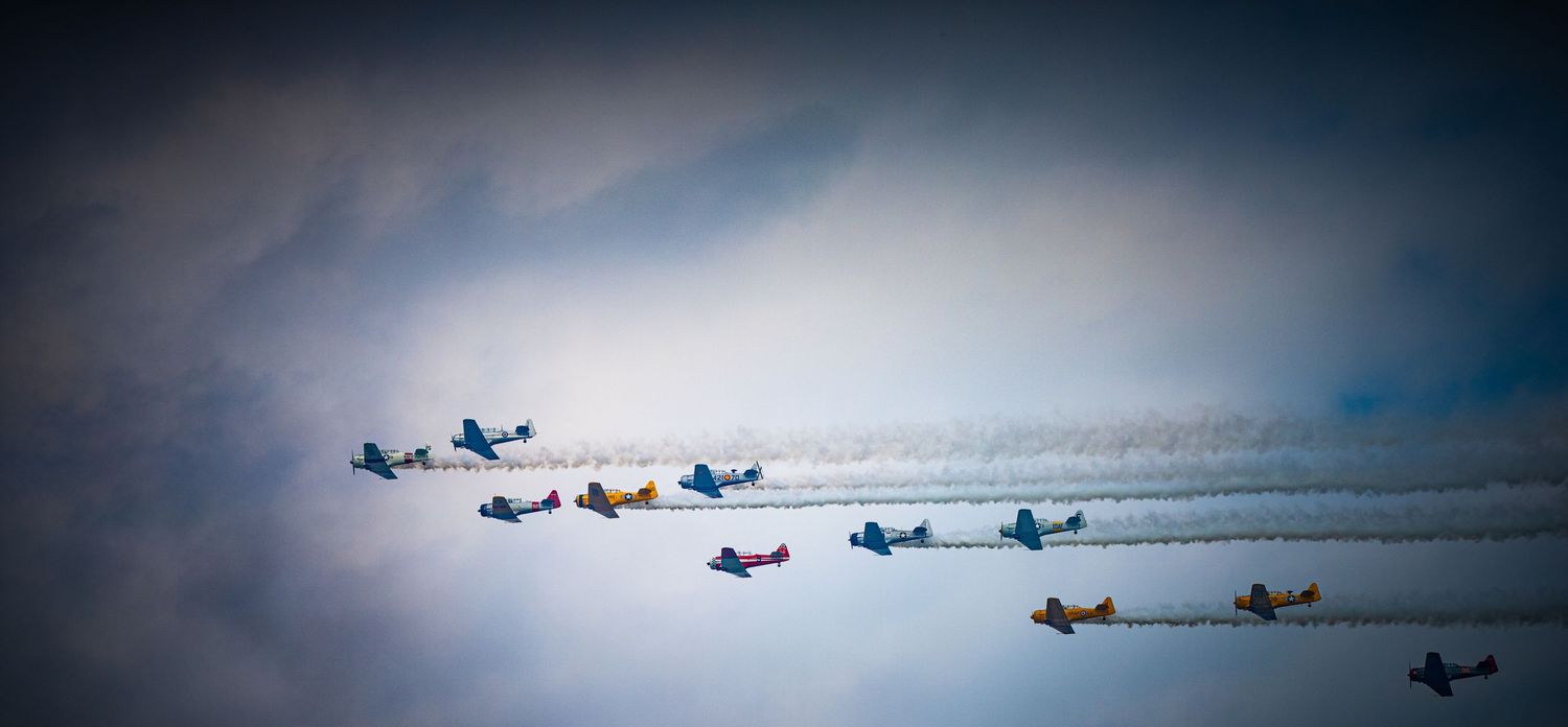 Formation of vintage aircraft flying through cloudy skies leaving contrails behind.
