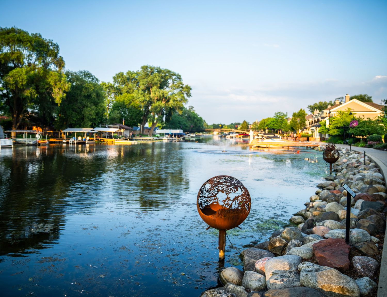 Waterfront path with decorative metal orbs and rock barriers along a calm lake.