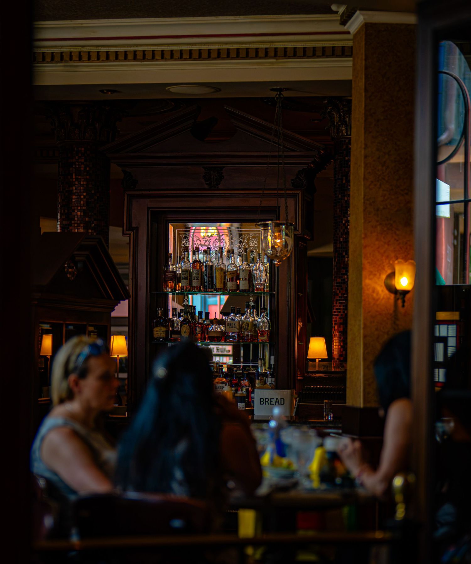 Dimly lit bar interior with patrons seated at tables, warm lamp glow, and a well-stocked liquor shelf visible through a doorway.