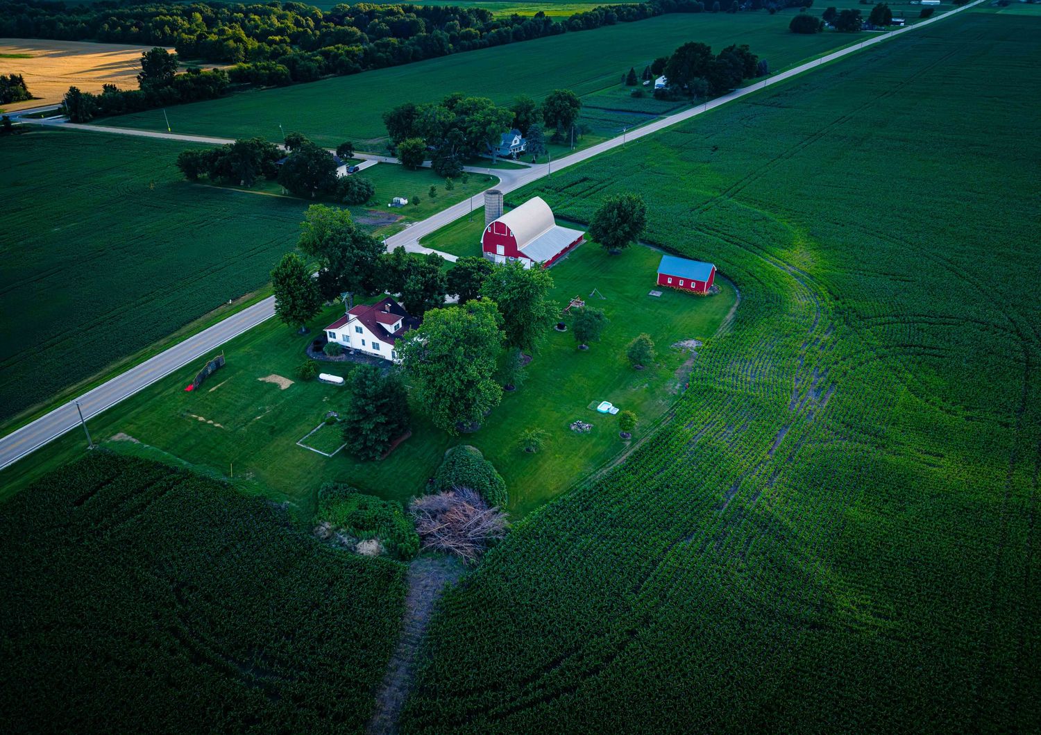 Aerial view of a rural farm with red barns, white farmhouse, and lush green crop fields along a country road.