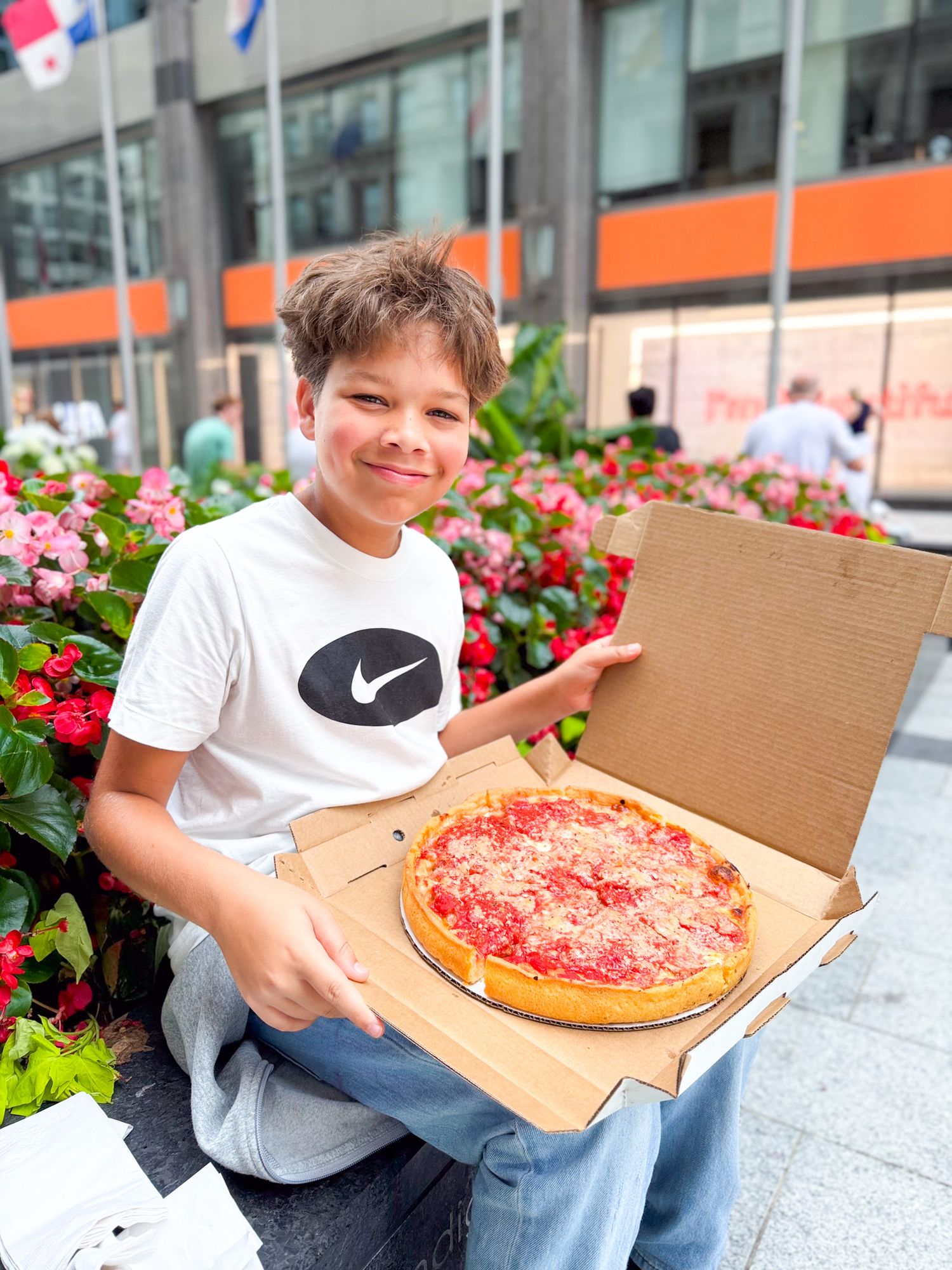 Boy in Nike shirt smiling while holding an open pizza box with a deep dish pizza outdoors near flowers.