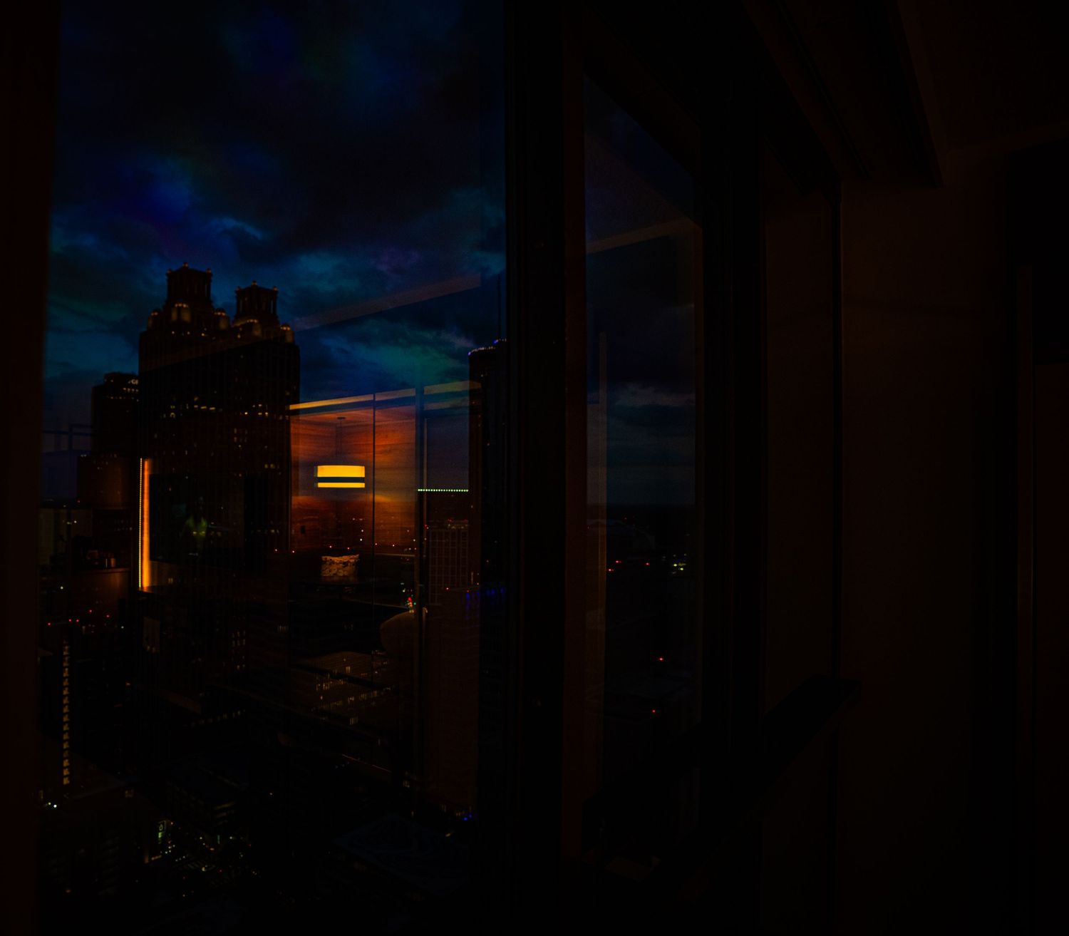 Dark cityscape view through a window at night with glowing lights and dramatic blue cloudy sky.