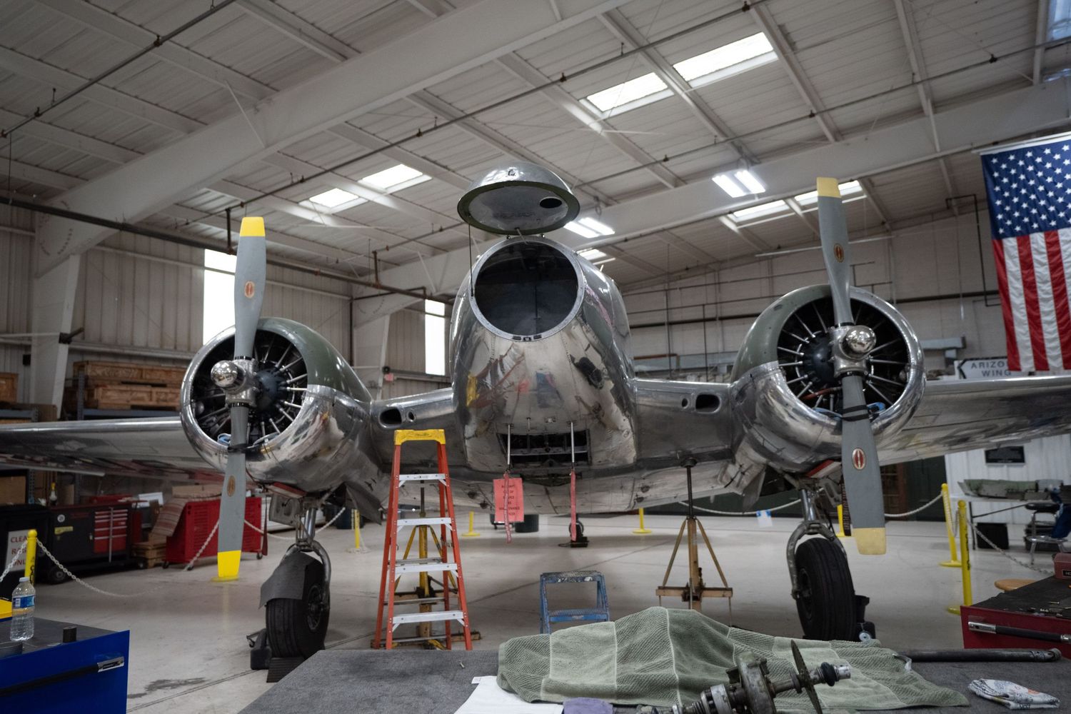 Vintage twin-engine aircraft undergoing restoration in a hangar with an American flag displayed on the wall.