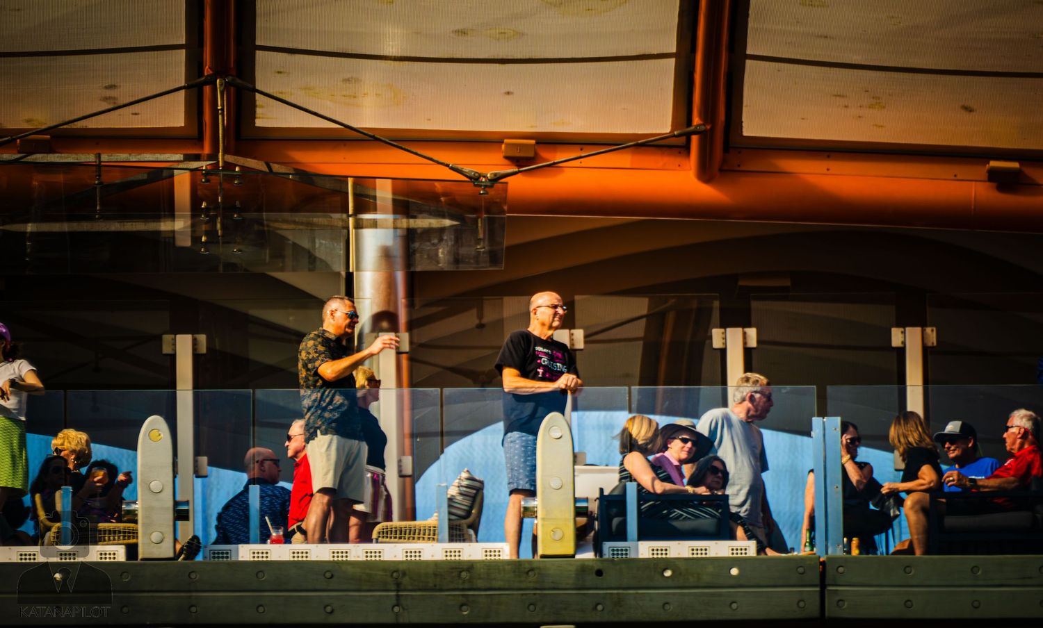 Silhouettes of people waiting in line against an orange sunset sky at a fairground or outdoor event.
