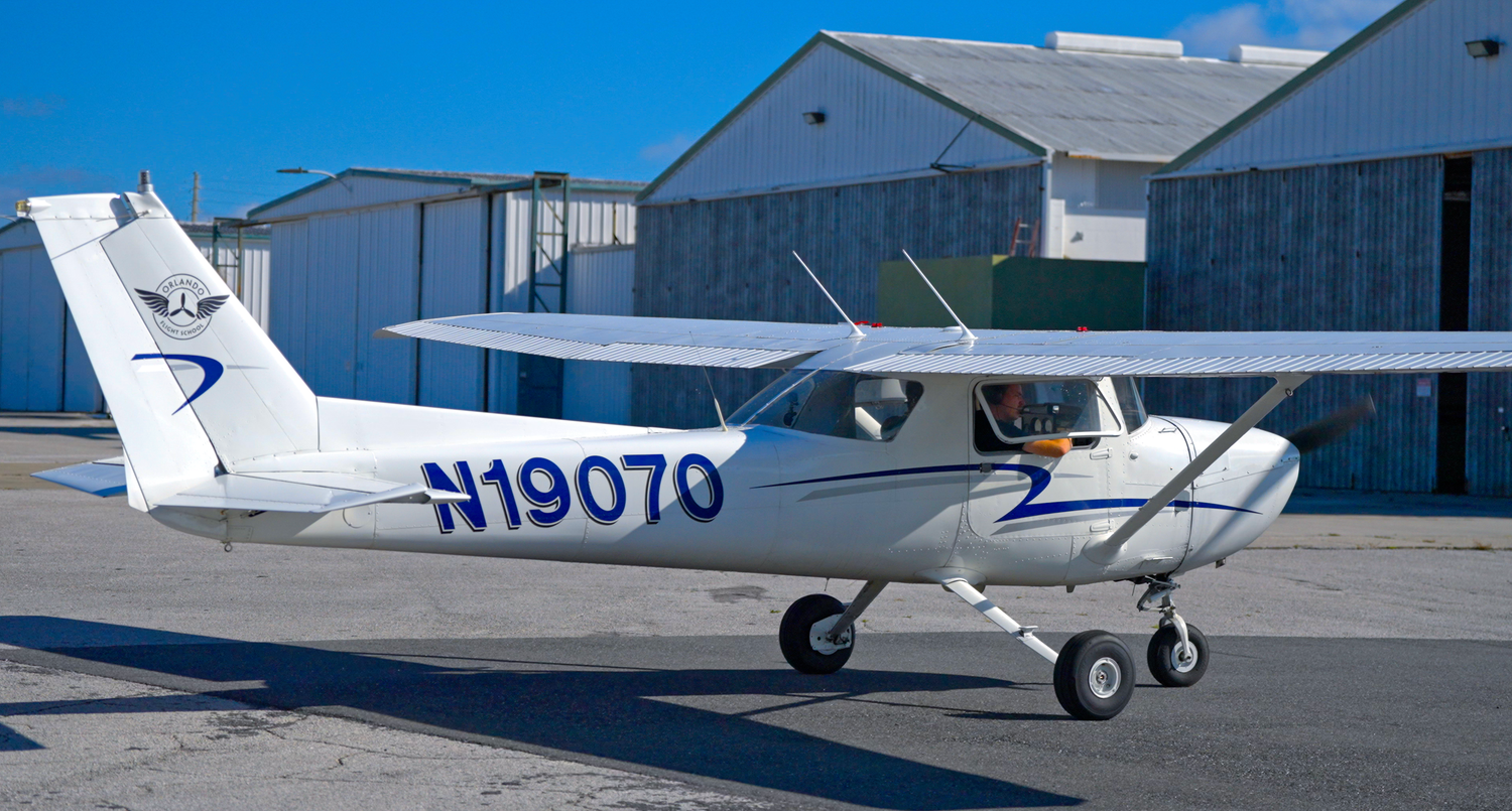 Row of small aircraft parked outside hangars at an airfield under blue sky.